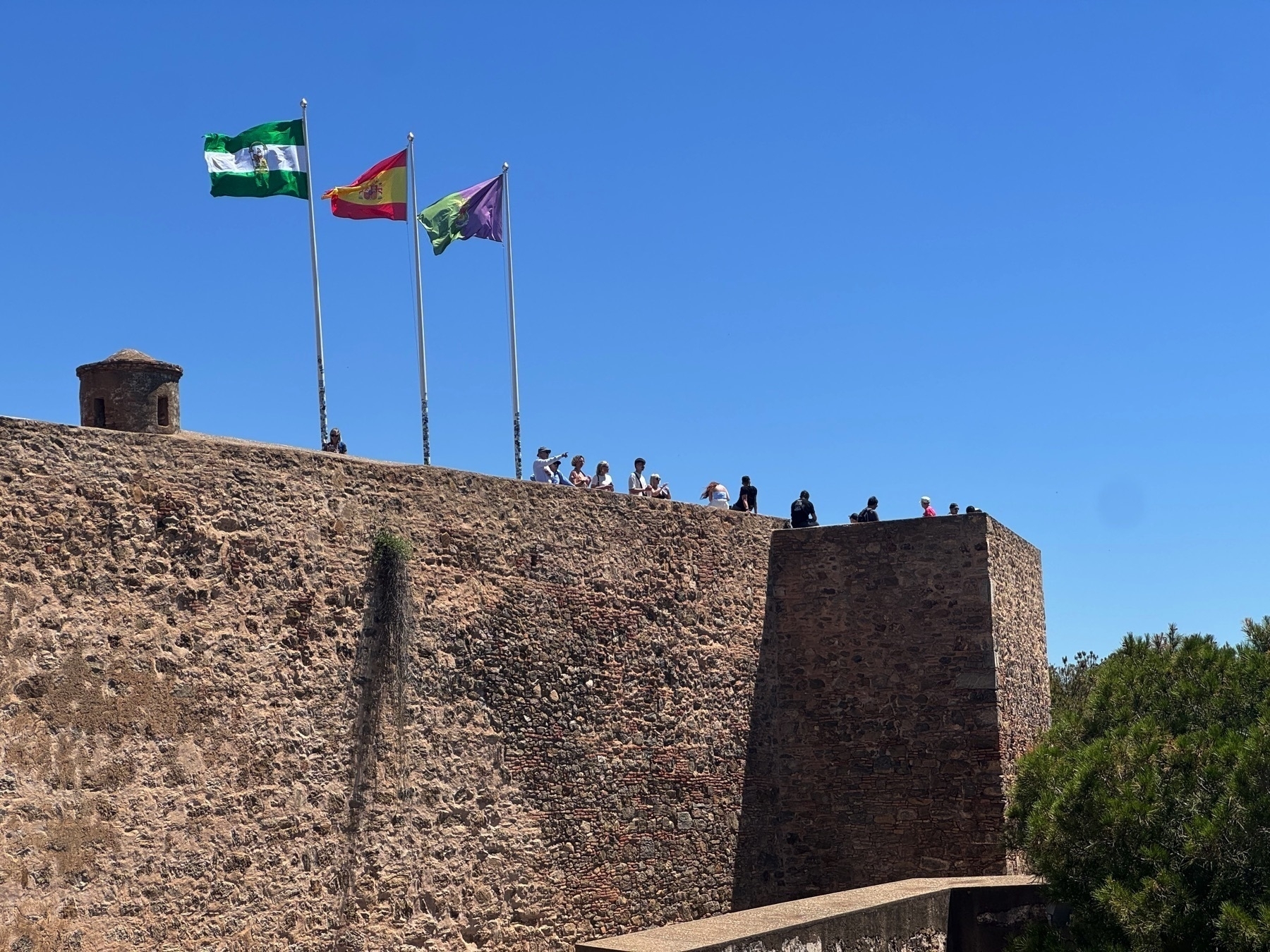 Tourists gather along a high stone rampart under three waving flags—Andalusia, Spain, and Málaga—on a clear sunny day. A small turret stands at the corner, evoking the structure’s defensive history.