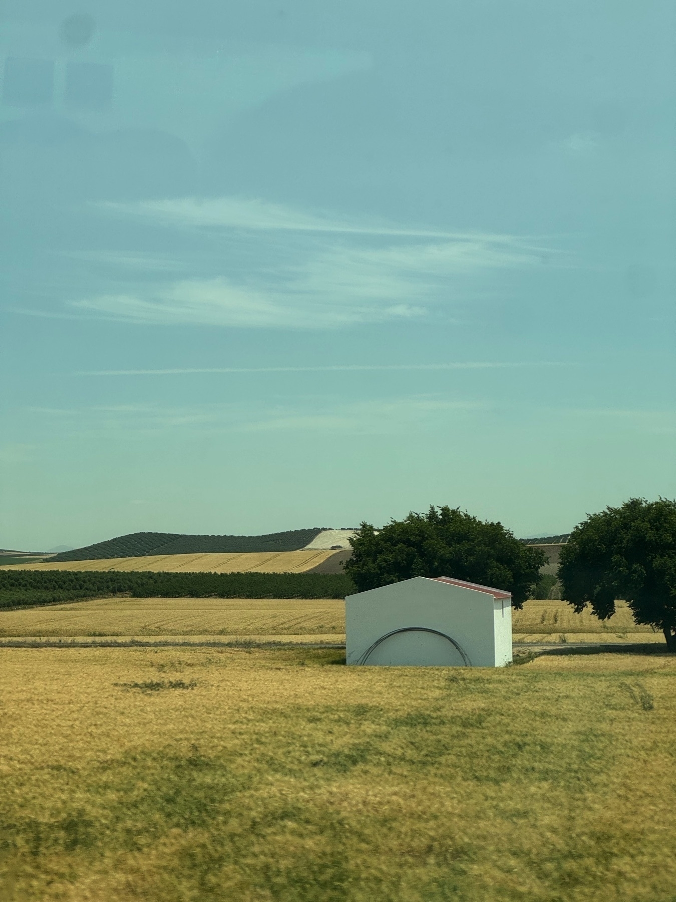 A small white utility structure with a curved detail on its front stands in a golden field of dry grass. Behind it, rows of trees and patchwork farmland stretch across rolling hills under a pale blue sky with wispy clouds. Two large trees flank the structure, adding contrast and framing the rural scene with natural balance.