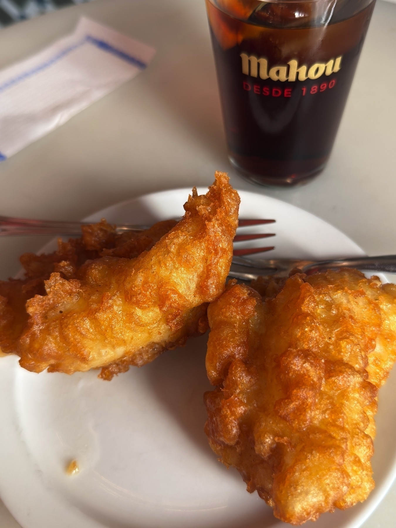 Two thick, golden pieces of battered cod served on a plain white plate, their crunchy exterior glowing in the sunlight. The drink beside them, a dark soda in a glass branded with “Mahou”, adds a local touch and refreshing contrast to the hearty fried fish.