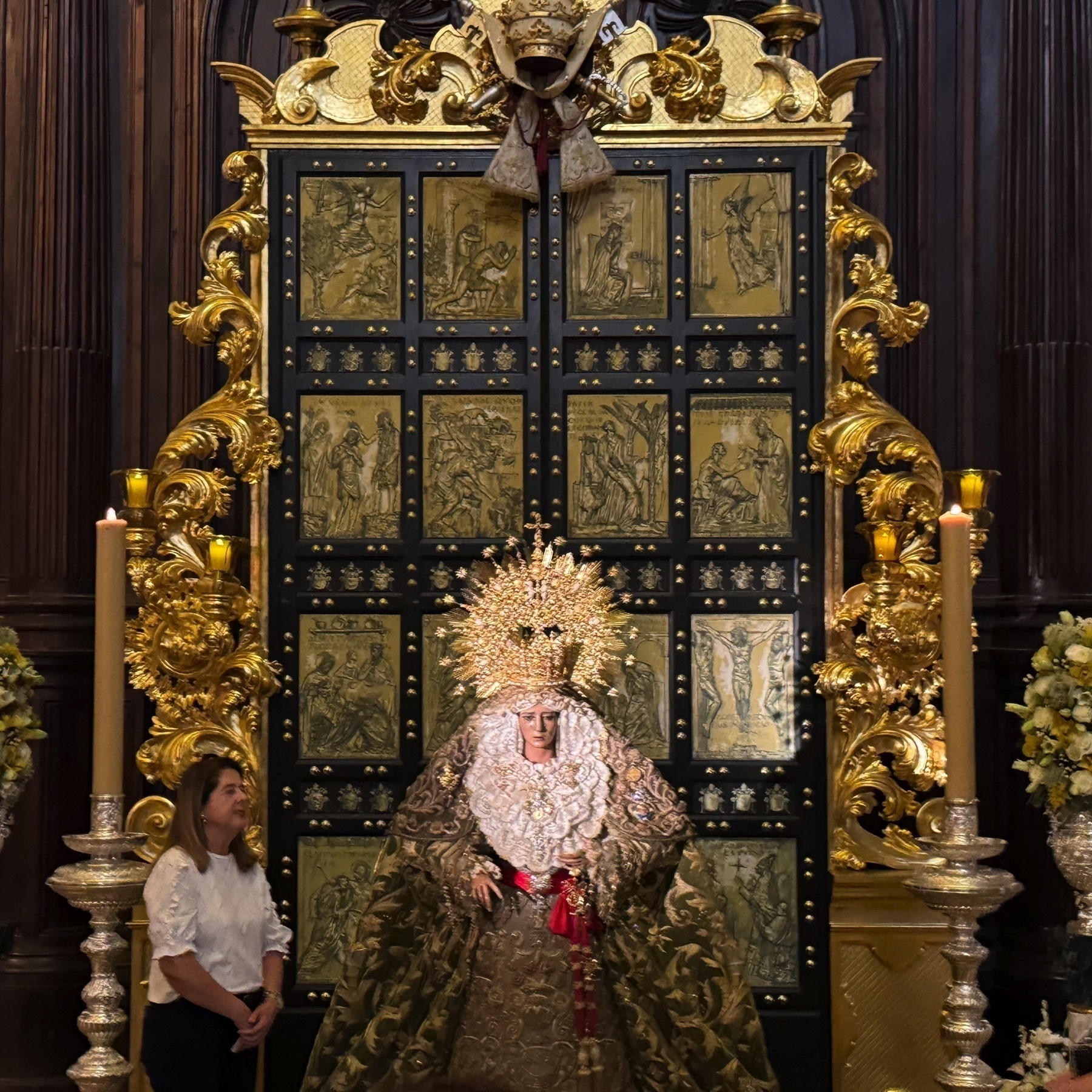 A statue of the Virgin Mary dressed in a richly embroidered robe and an ornate gold crown, flanked by large golden candle holders and flower arrangements. The black and gold backdrop features engraved religious scenes, contributing to a solemn and sacred atmosphere as visitors look on.