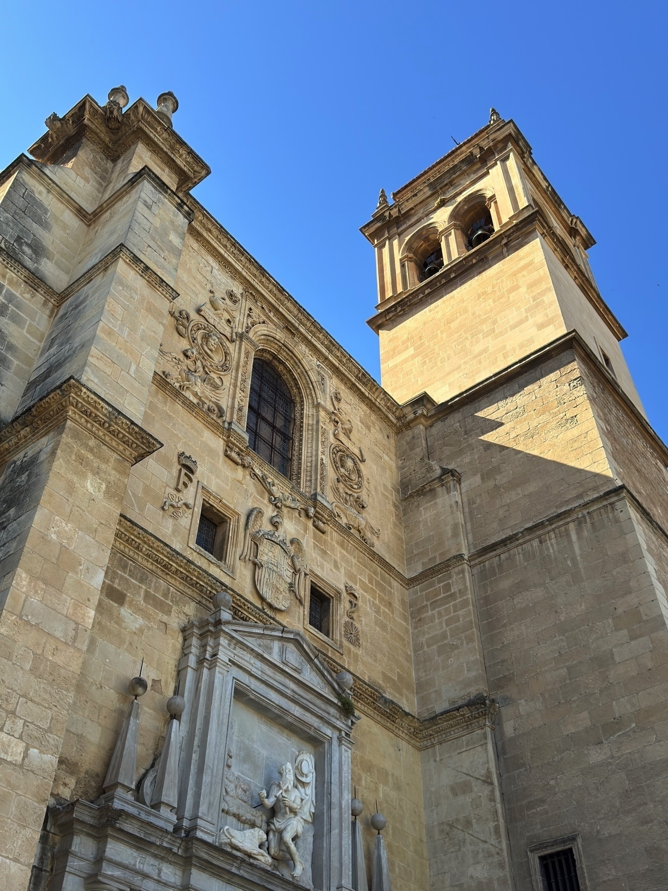 Upward perspective of the stone bell tower and ornate facade, seen against a clear blue sky. The structure features Renaissance-style carvings, a sculpted relief above the main entrance, and detailed coats of arms, emphasizing the building’s historical and architectural richness.