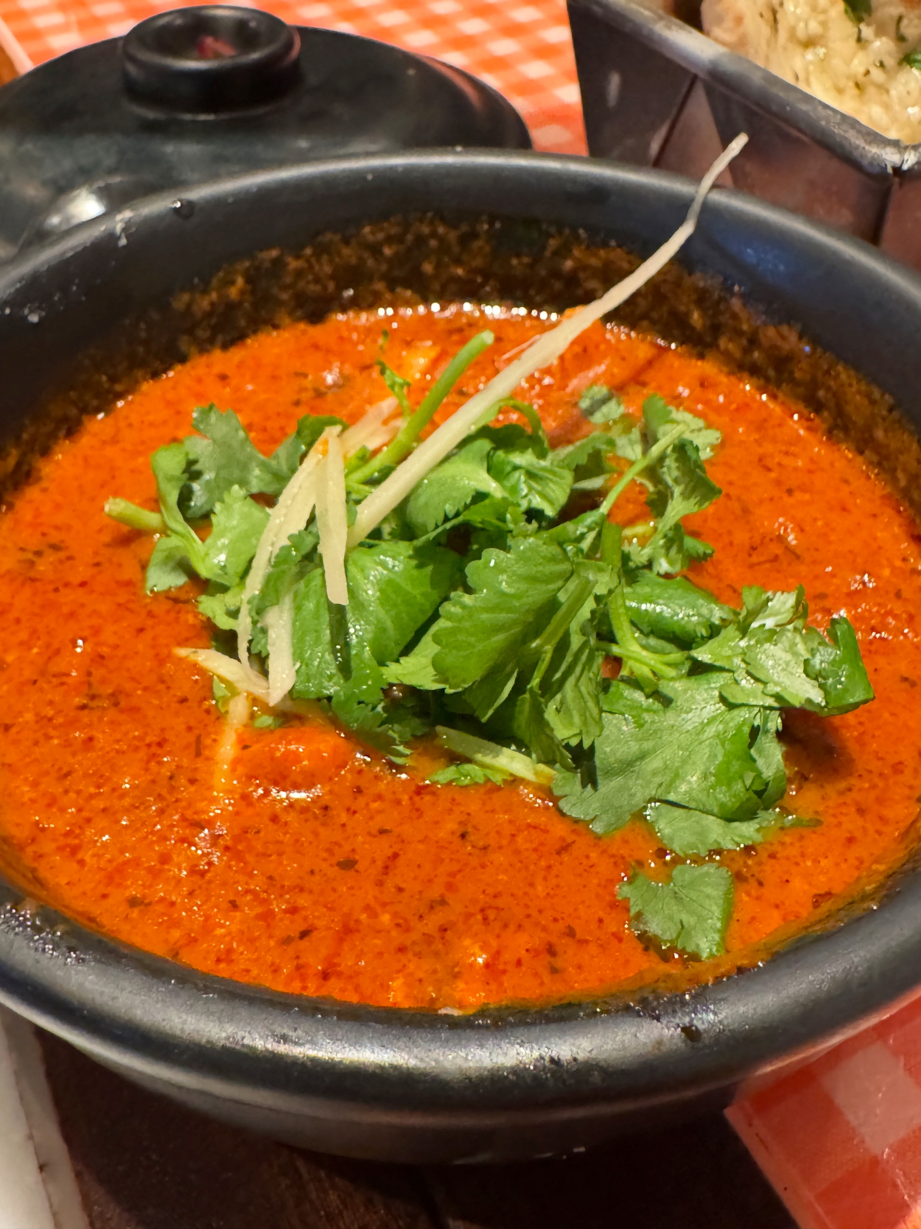 A bowl of vivid red curry with a glossy surface is topped with fresh coriander and thin strips of ginger. The oil-speckled sauce looks rich and warming, with the bright green garnish adding contrast and freshness to the deeply spiced base.