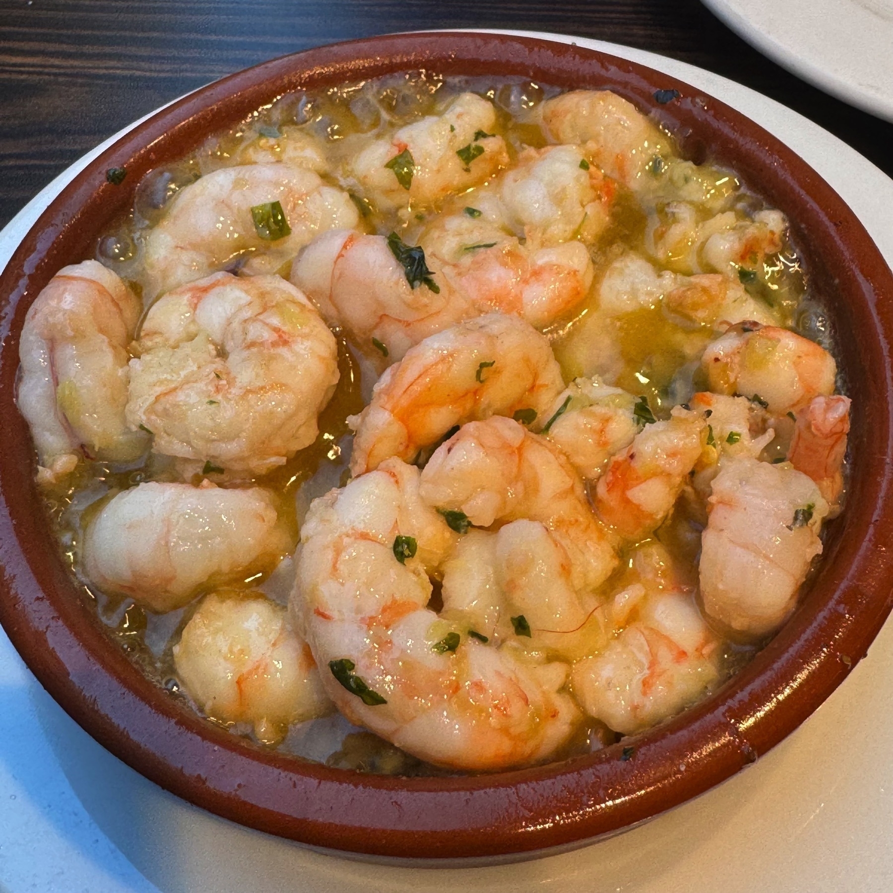 A terra-cotta dish filled with sizzling garlic shrimp swimming in bubbling olive oil, speckled with bits of fresh parsley. The shrimp appear plump and juicy, their slight char and vibrant color enhancing the sense of flavor and heat.