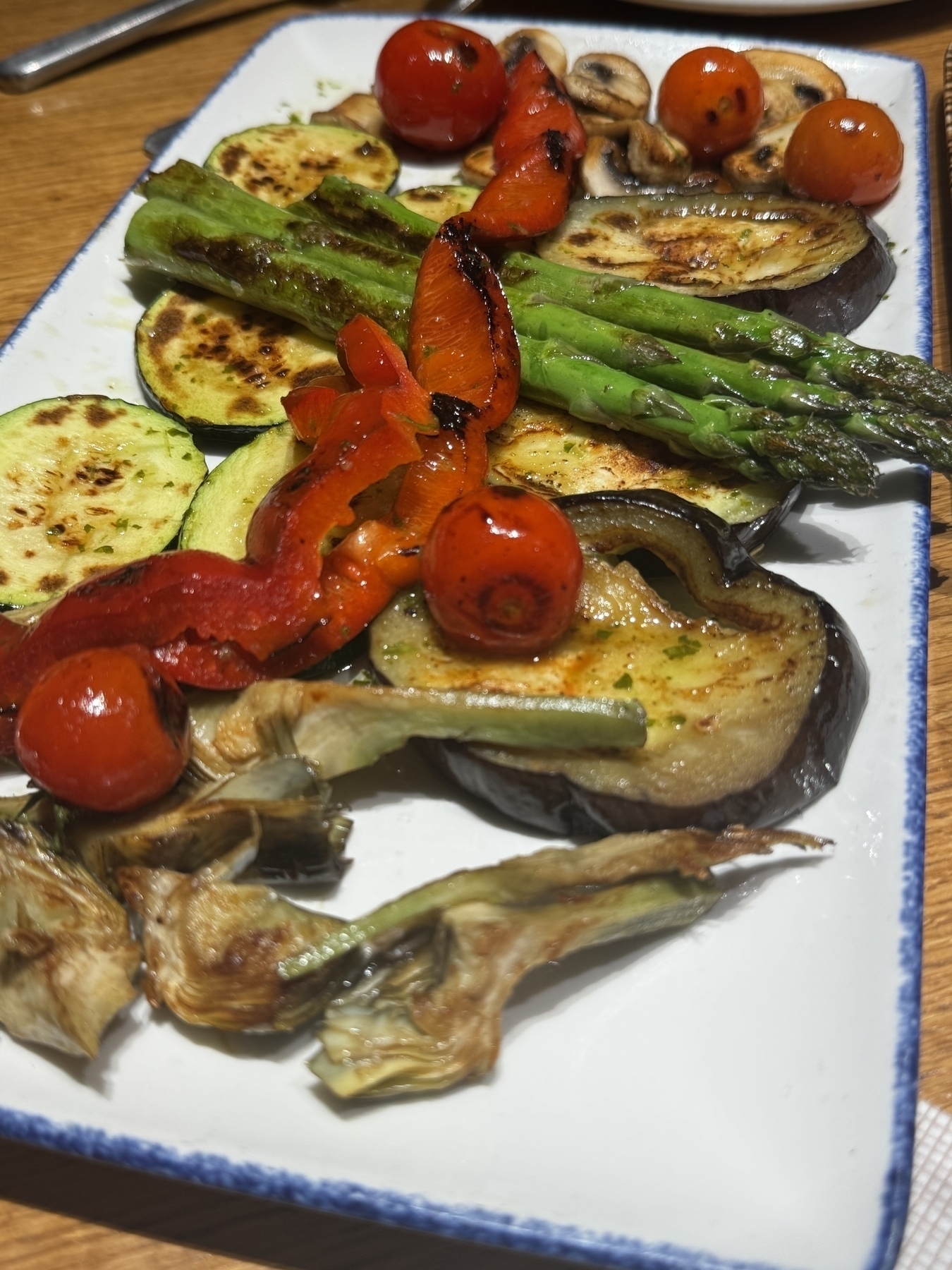 A platter of grilled vegetables including zucchini, eggplant, asparagus, red pepper, mushrooms, and cherry tomatoes is arranged on a rectangular plate.
