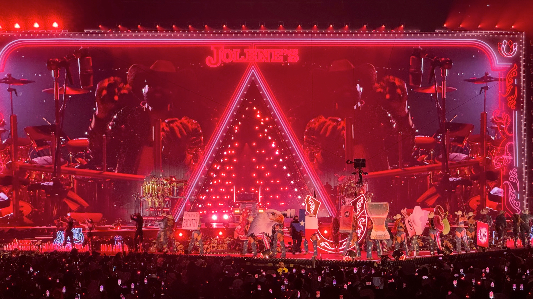 The stage glows deep red as Beyoncé performs “Jolene,” surrounded by a theatrical Western saloon–inspired set. A large neon sign reading “Jolene’s” crowns the stage above a triangular light structure, while dancers in cowboy-themed costumes and bold props move across the stage, creating a fiery, high-drama atmosphere.