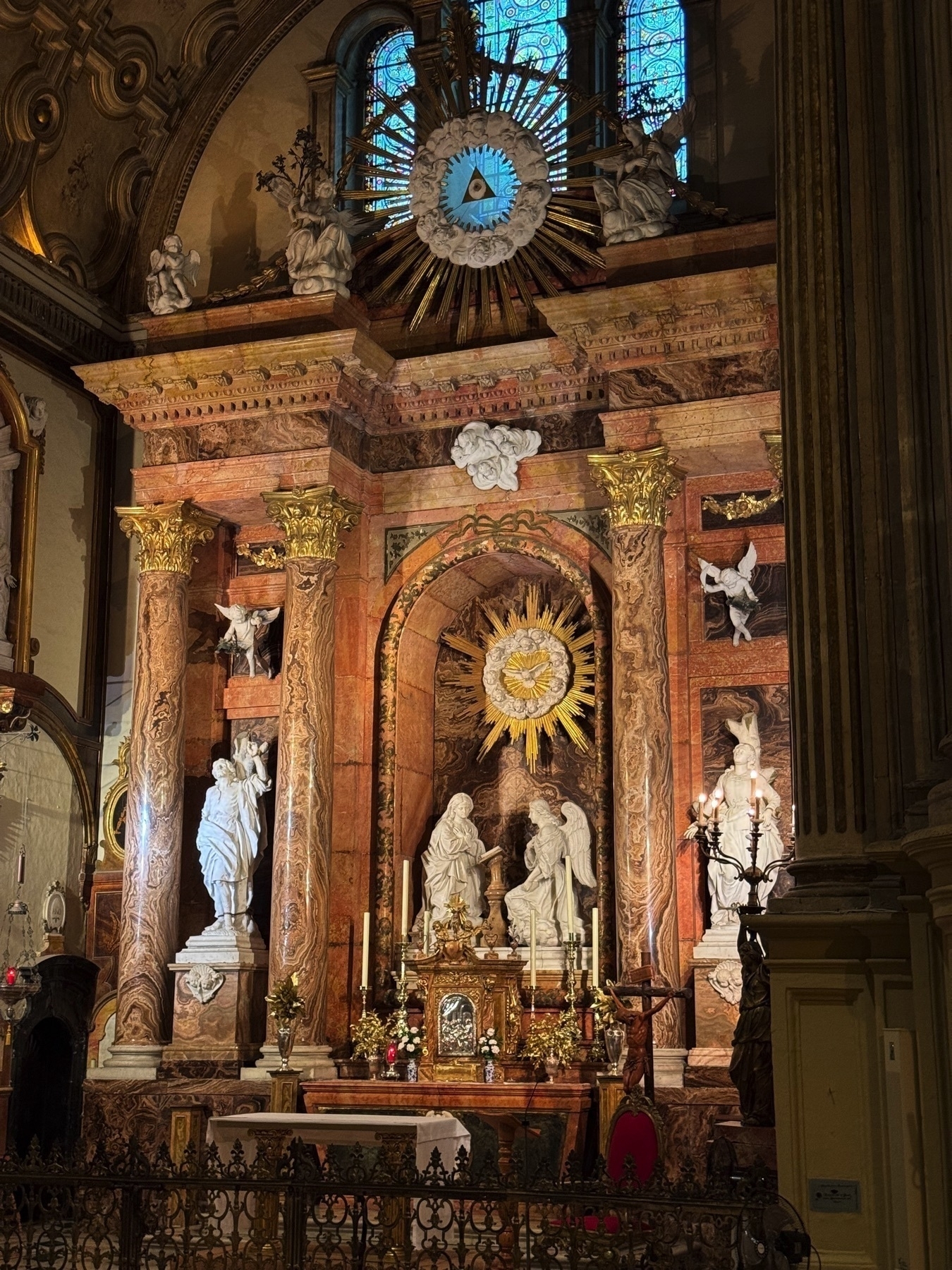 A richly decorated altar featuring pink marble columns, white statues of angels and saints, and golden rays radiating from a triangle symbol representing the Holy Trinity. The stained glass in the background filters light, enhancing the warmth of the scene.