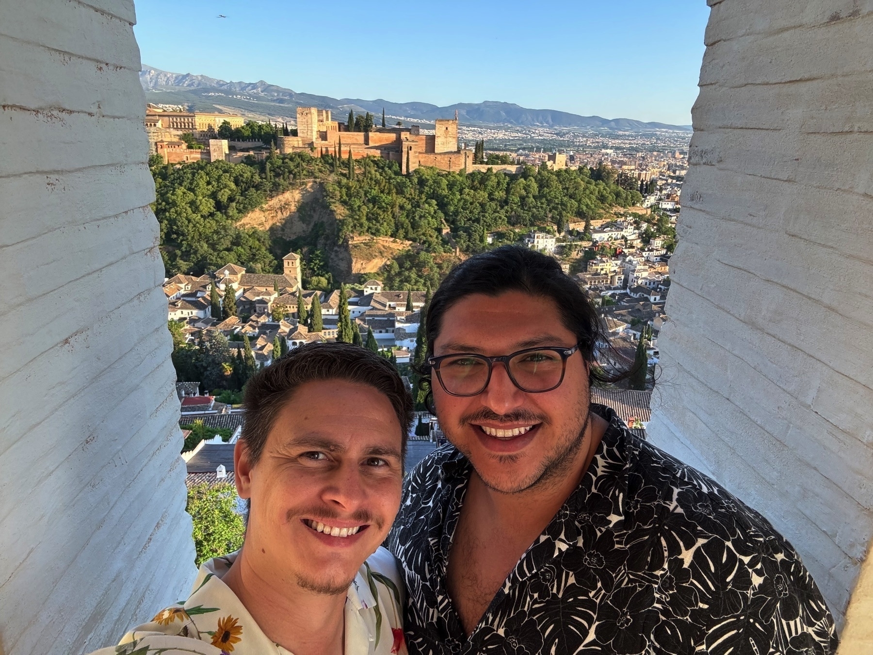 Two smiling people posing between whitewashed walls with a stunning panoramic view of the Alhambra in the background, perched on a lush hill above the city of Granada. The late afternoon sunlight casts a golden hue over the fortress and surrounding town, while mountains stretch along the horizon.