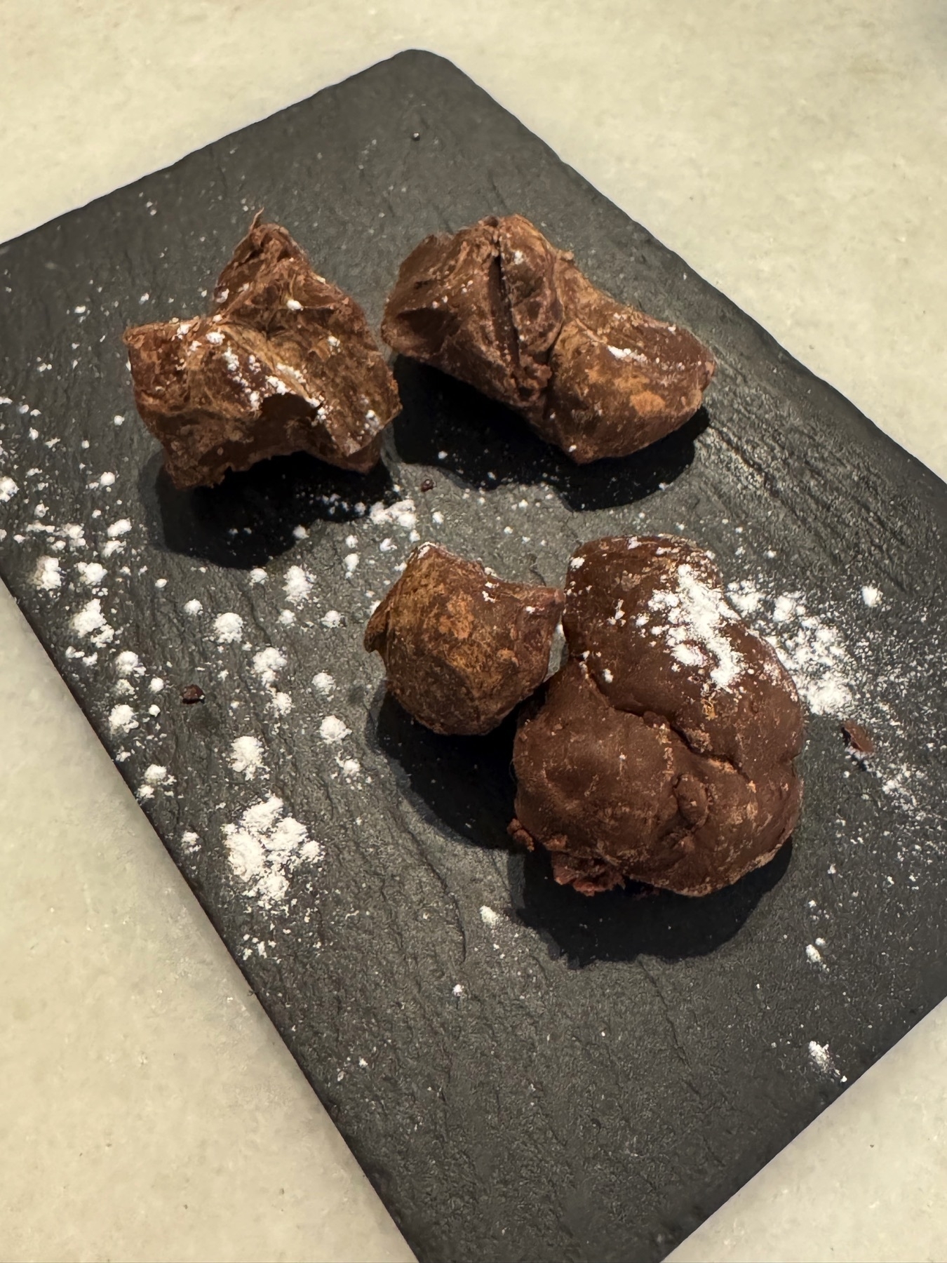 Three hand-formed chocolate truffles with a rough, rustic finish, dusted lightly with powdered sugar and arranged on a matte black slate. The cracked surfaces and rich brown color suggest a dense, fudgy interior, offering an indulgent, artisanal dessert experience.
