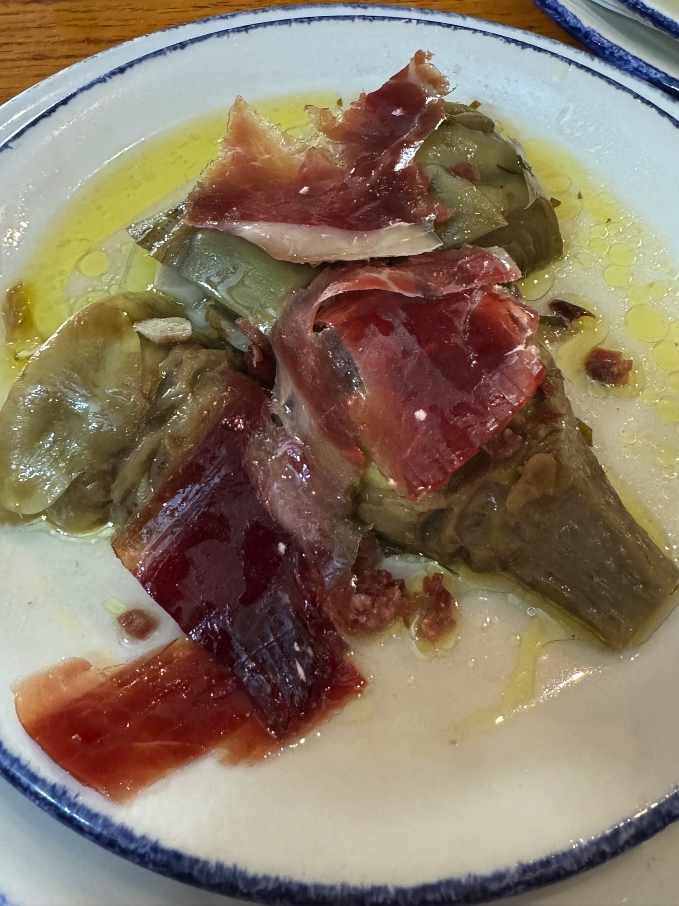 Tender artichoke hearts bathed in olive oil and topped with silky slices of jamón ibérico, creating a luscious contrast between the delicate vegetable and the savory, cured ham. The oil pools on the plate, adding a glossy finish that makes the dish look indulgent and rich.