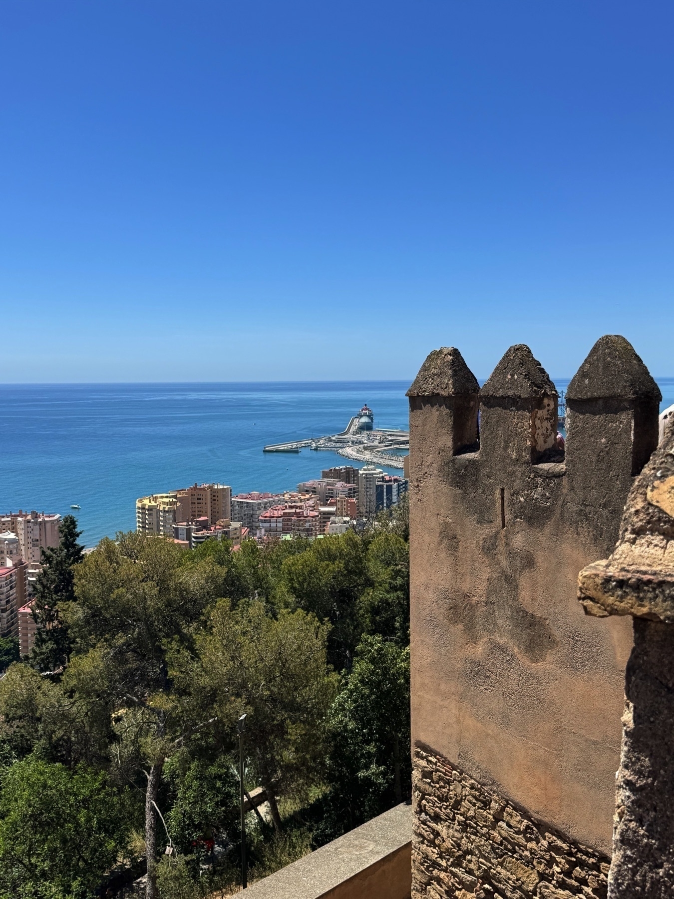Coastal view from a high vantage point showing a stone turret in the foreground and a lighthouse at the tip of a long breakwater in the distance. Below, high-rise buildings border the sea, framed by lush green trees.
