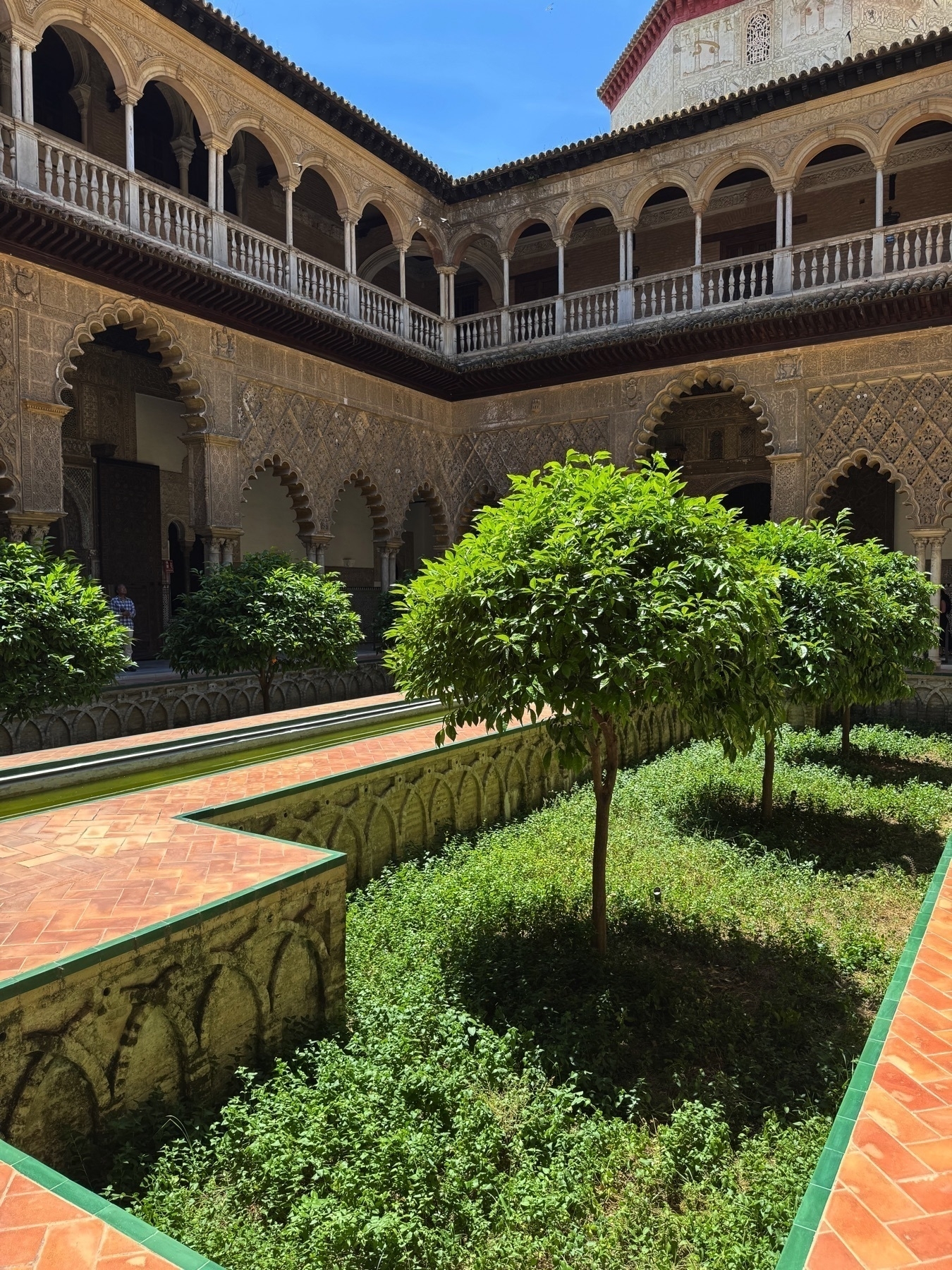 The Courtyard of the Maidens, with orange trees planted in sunken garden beds and a narrow central reflecting pool. The surrounding Mudéjar arcade features scalloped arches with intricate stucco work and a second level of Gothic-style arches supported by slender columns.