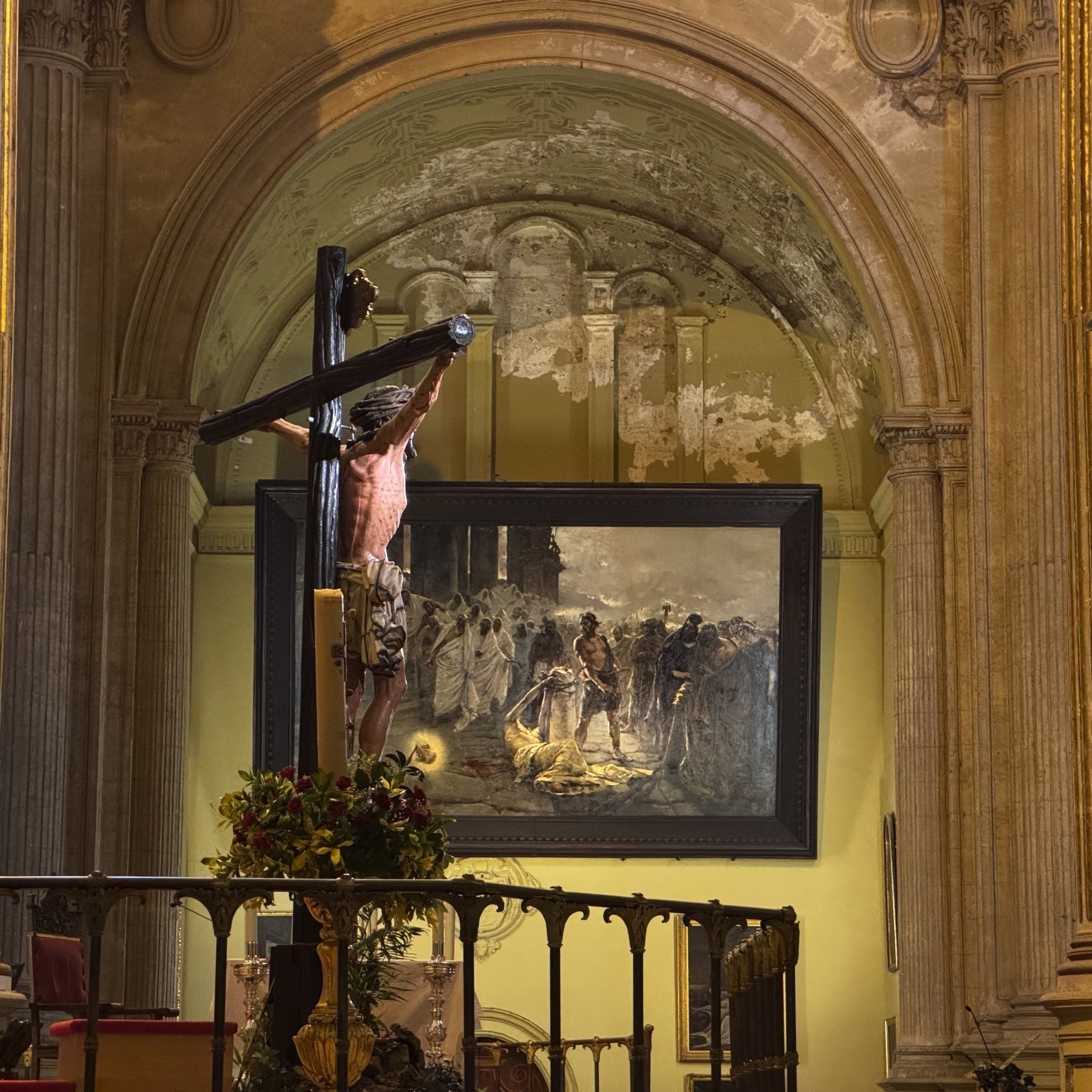 A wooden crucifix of Christ on the cross positioned in front of a dramatic painting depicting a somber biblical scene. The surrounding arch is weathered, revealing patches of exposed stonework that contrast with the grandeur of the rest of the cathedral.