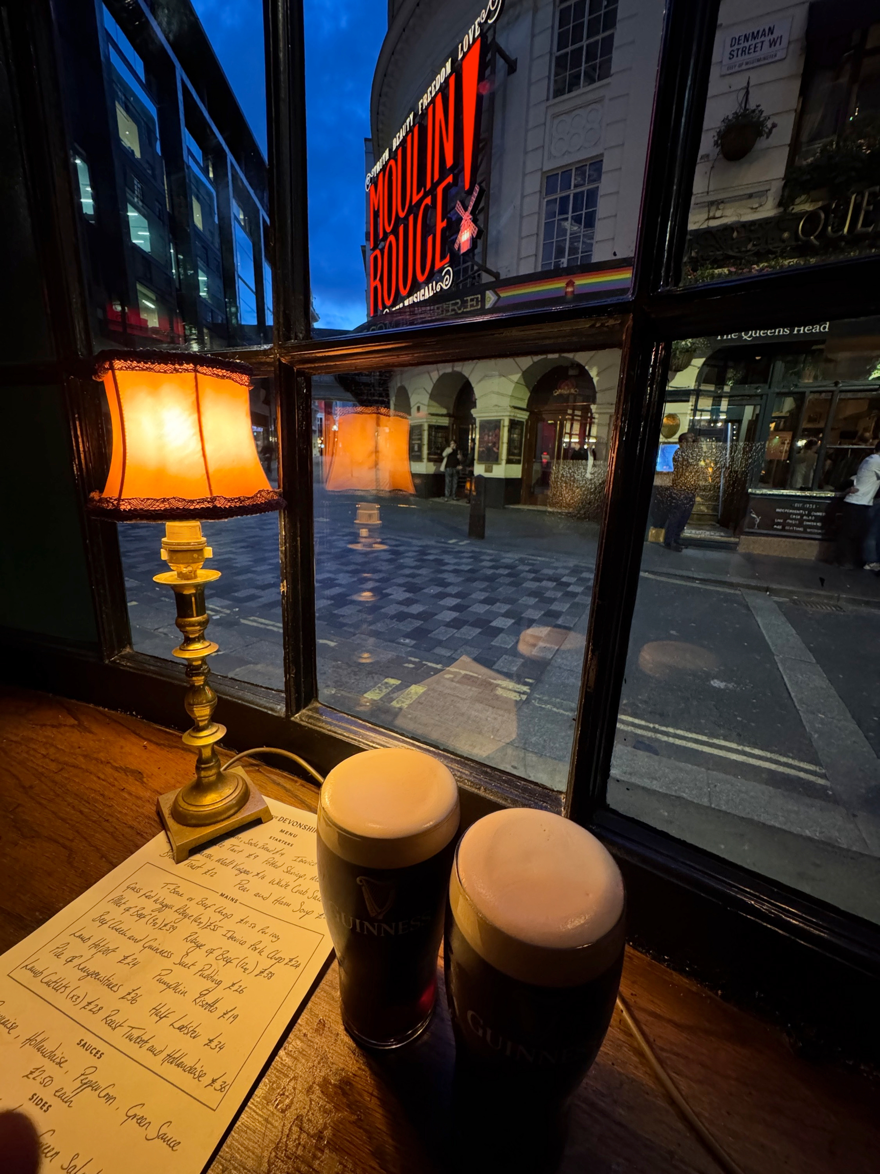 Two pints of Guinness rest on a wooden window ledge beside a small table lamp and a handwritten menu, while the Moulin Rouge sign shines outside through the glass, mixing pub warmth with busy West End night energy.