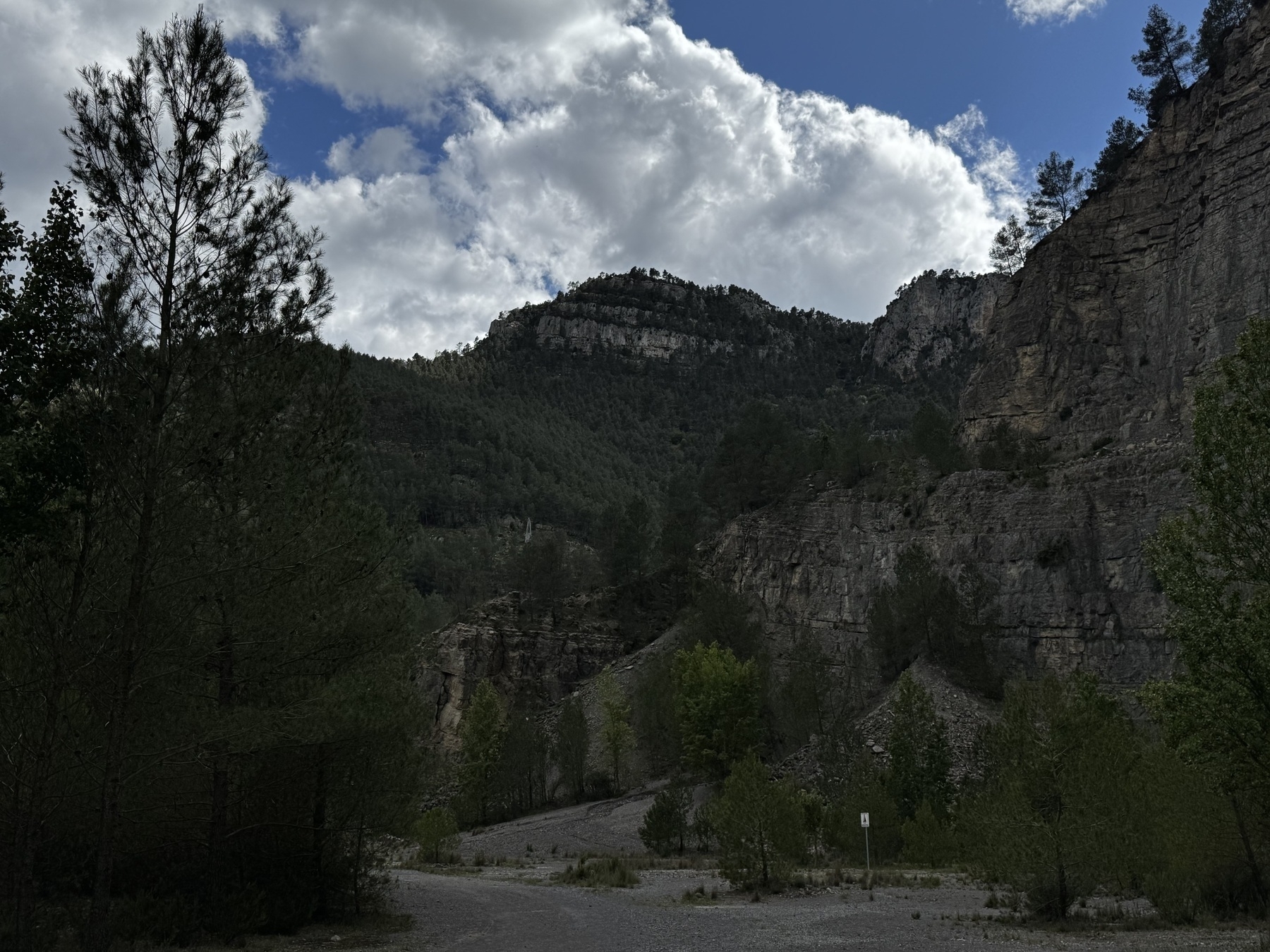 A rugged mountain landscape with cloudy skies above and steep, forested hills enclosing a dirt trail at the bottom of the valley.