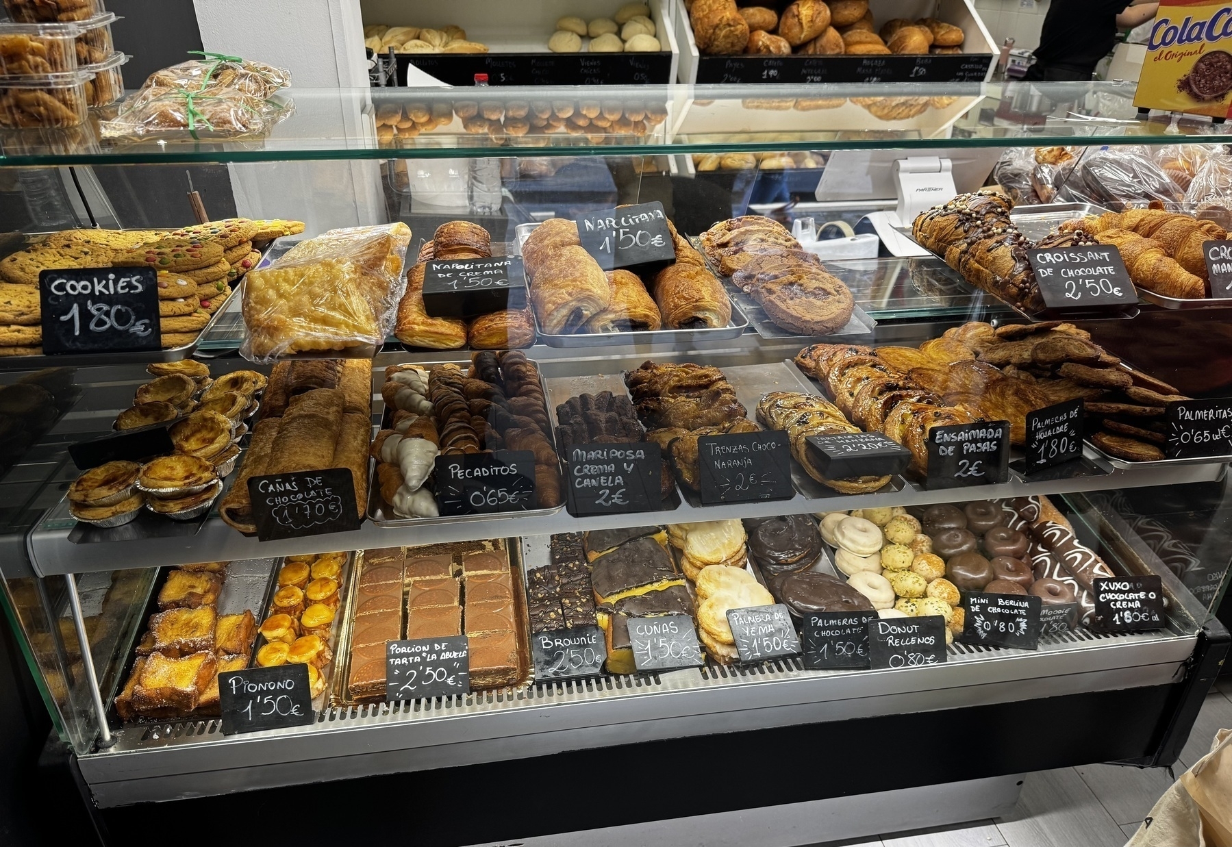 Wide shot of a glass display case at Panadería Ana filled with a variety of baked goods, including cookies, croissants, puff pastries, brownies, and chocolate-covered treats. The assortment is neatly labeled with handwritten price signs, and the diversity of shapes, colors, and textures—from flaky layers to glossy chocolate coatings—makes the case visually rich and inviting.