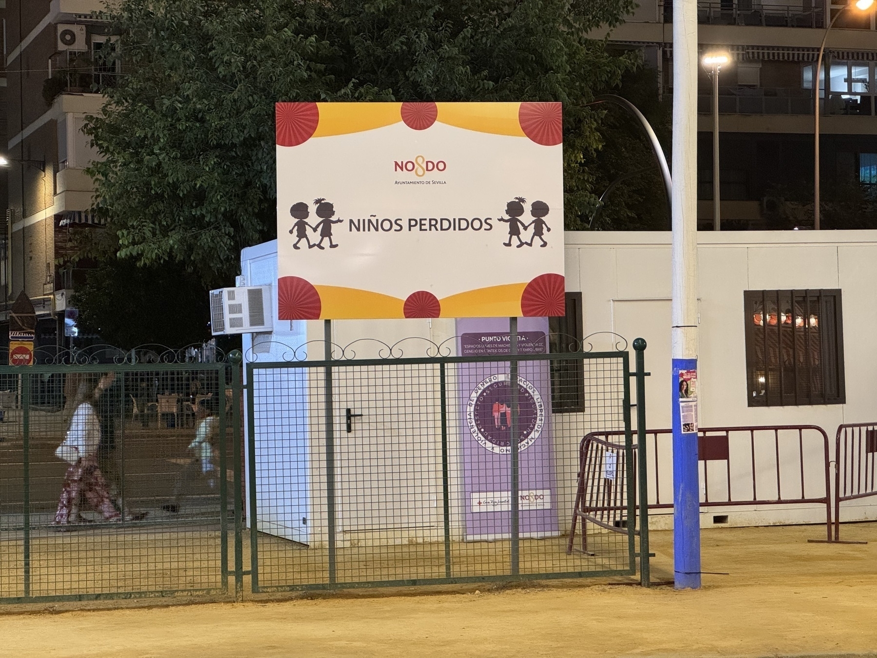 A small white building labeled “Niños Perdidos” (Lost Children) with a cartoon sign of children holding hands, enclosed by a green fence. The setup highlights safety provisions for families during the event.