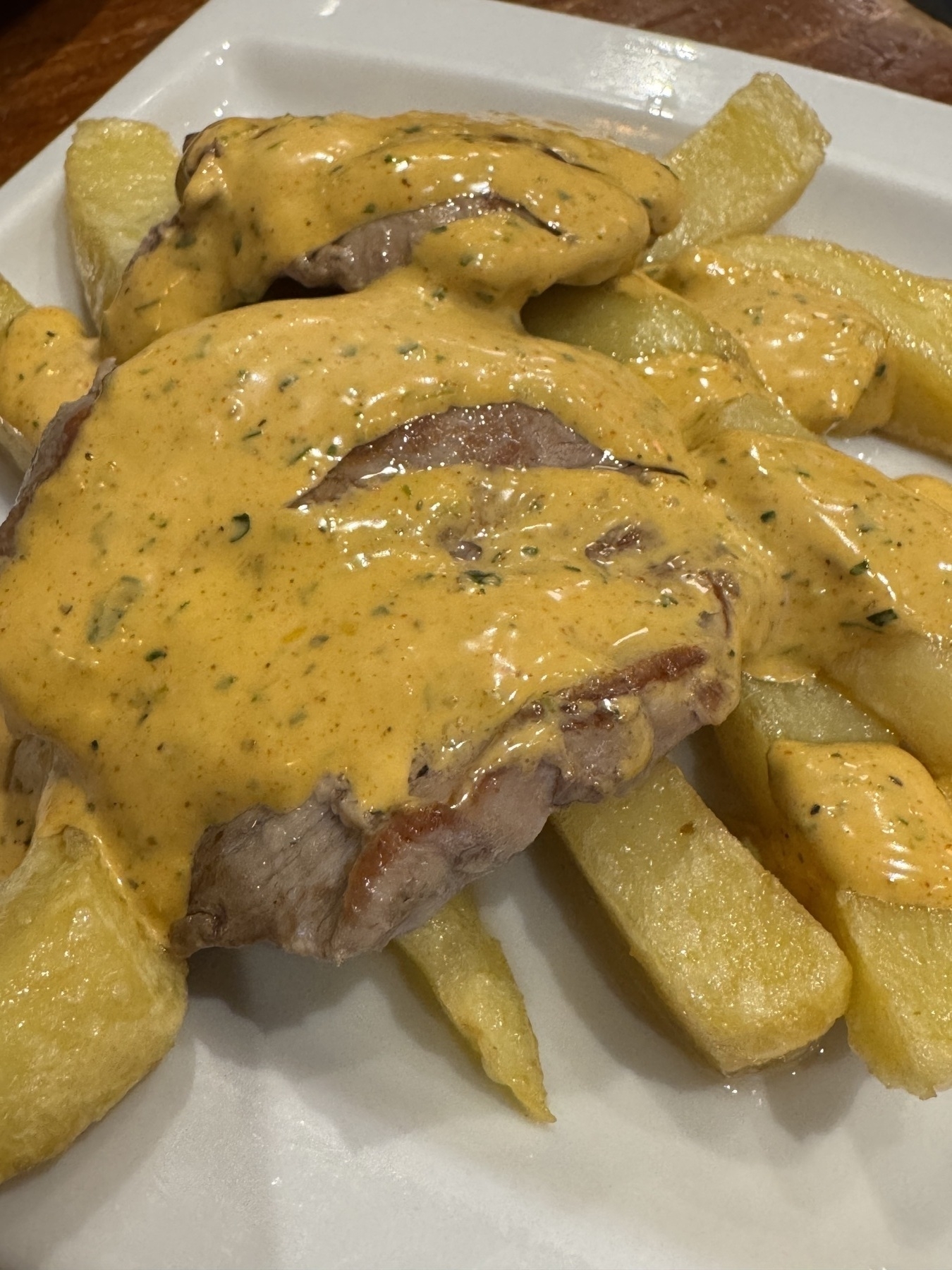 A white plate with Solomillo en salsa Mozárabe—grilled pork tenderloin medallions smothered in a creamy, orange-hued Moorish-style sauce. The dish is served with thick potato wedges. The warm color of the sauce and its speckled herbs evoke richness and aroma.