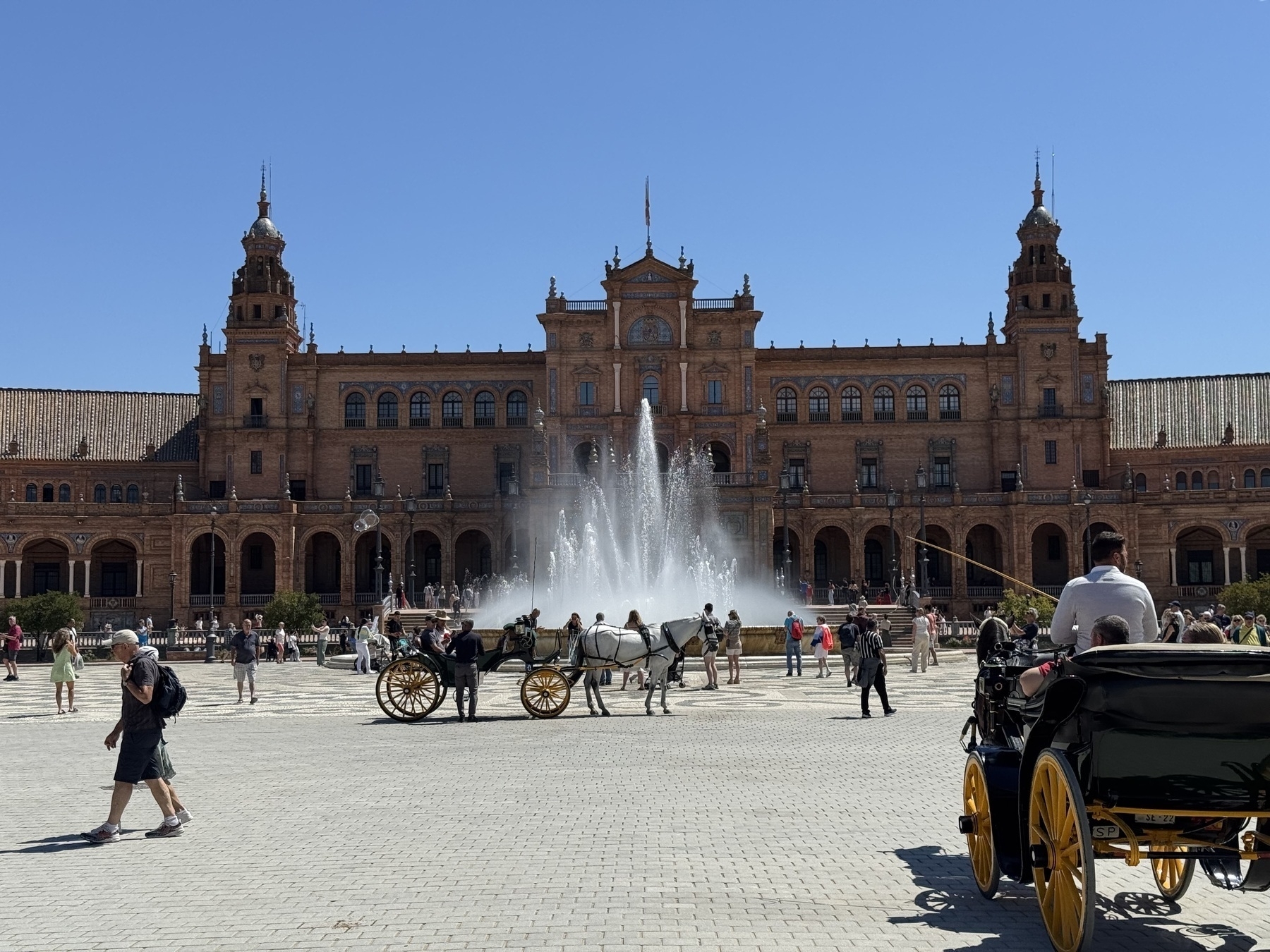 A bustling plaza scene with horse-drawn carriages circling a towering fountain, spraying water high into the air. Visitors stroll across the patterned square, backed by the grand central building with symmetrical towers and colonnaded galleries.