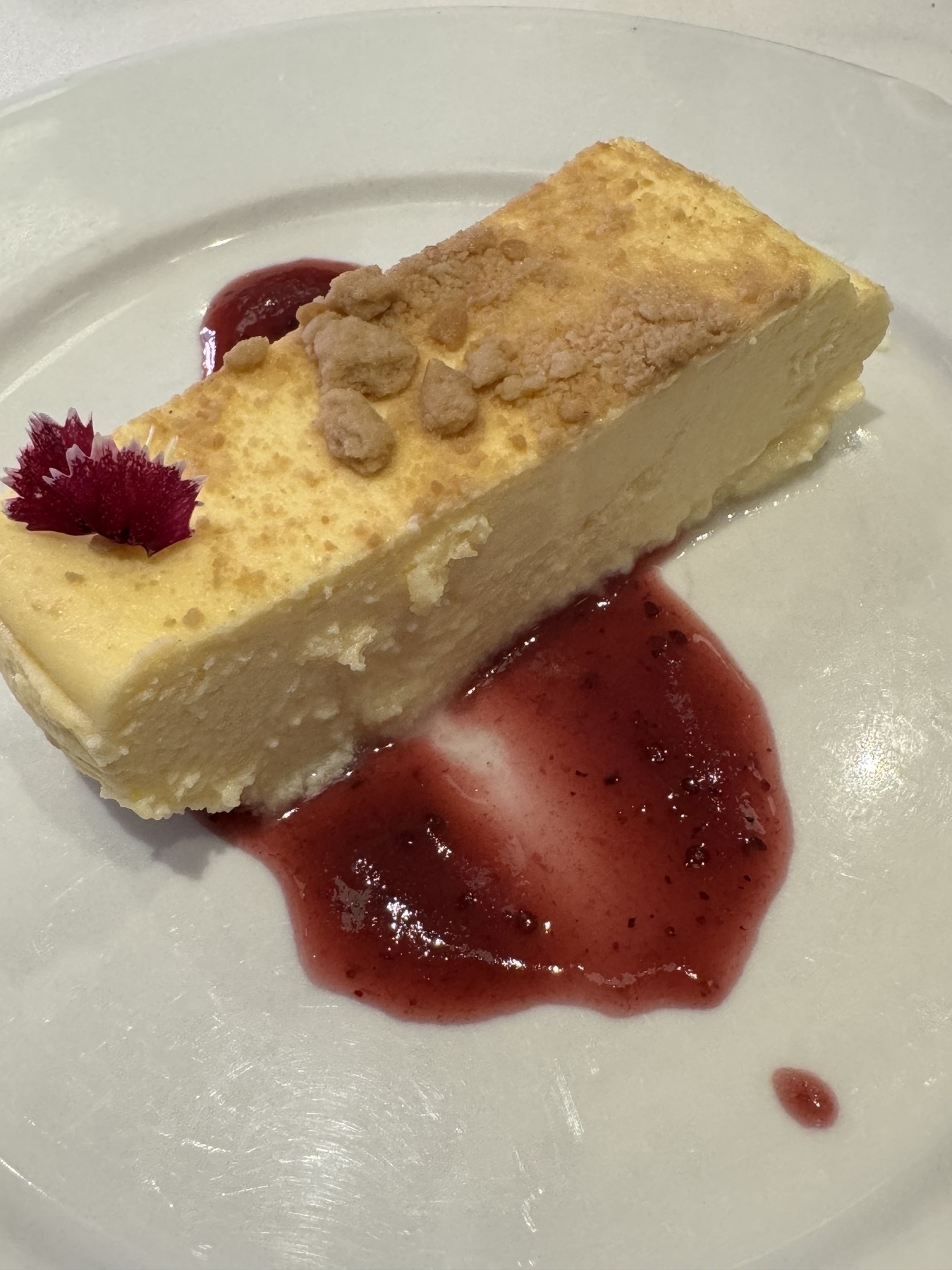 A slice of rich, baked cheesecake with a golden crumble topping, resting on a pool of glossy red berry sauce, garnished with an edible flower that adds elegance and a touch of color.