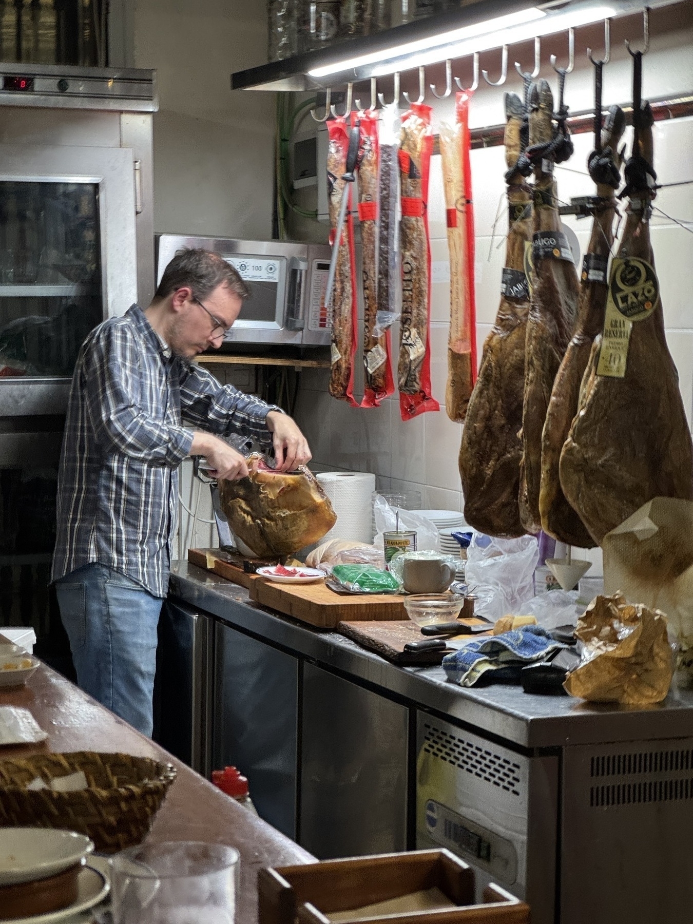 Man slicing a leg of jamón ibérico in a preparation area, surrounded by hanging cured meats and kitchen tools under bright lighting.