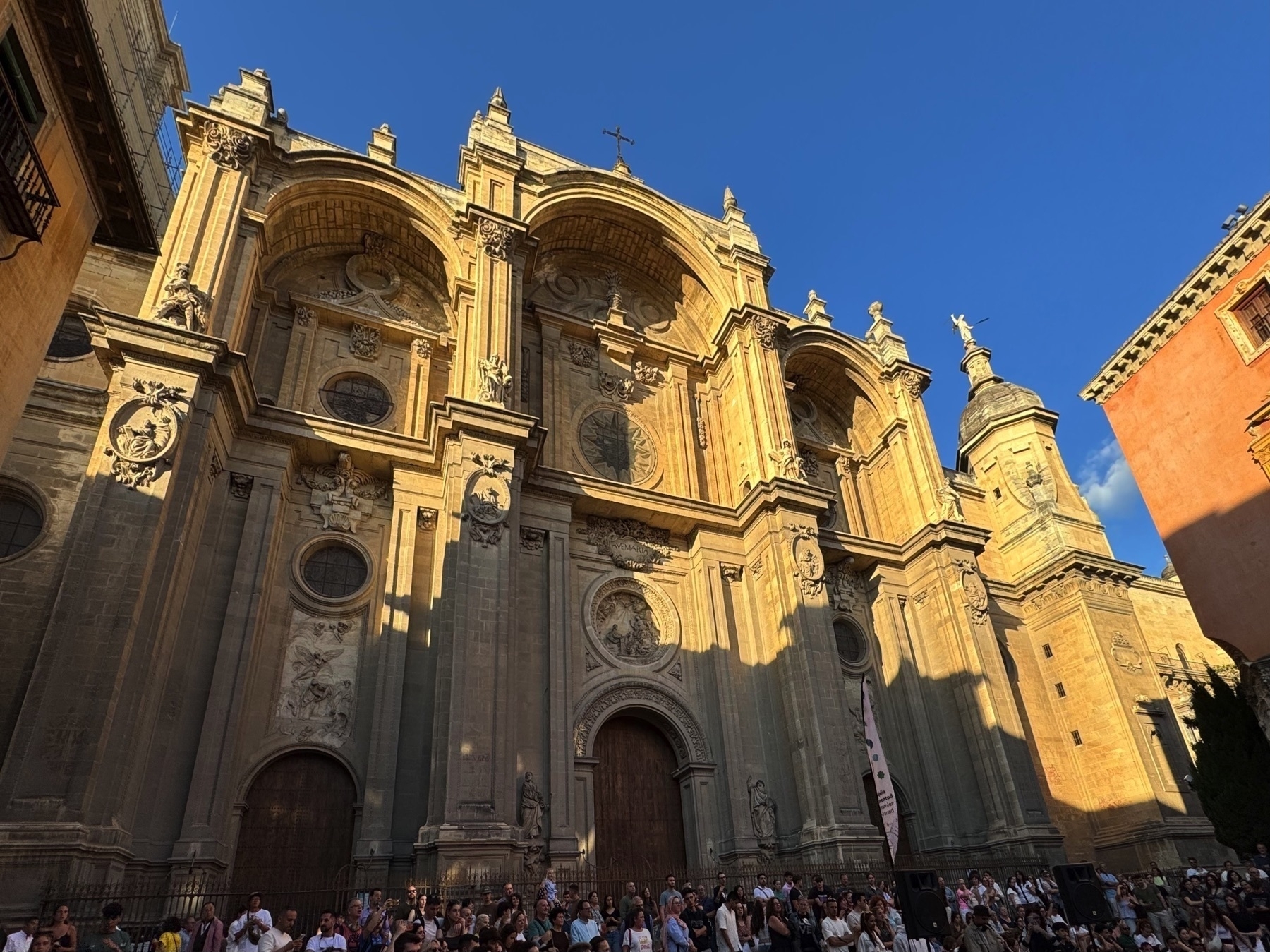 The grand façade of the Granada Cathedral under a clear blue sky, illuminated by the warm afternoon sun. The Renaissance-style exterior features soaring pilasters, elaborate sculptural reliefs, and three monumental arched entrances. Crowds gather along the base, suggesting a popular event or gathering. The strong sunlight casts dramatic shadows, emphasizing the depth and detail of the stonework.
