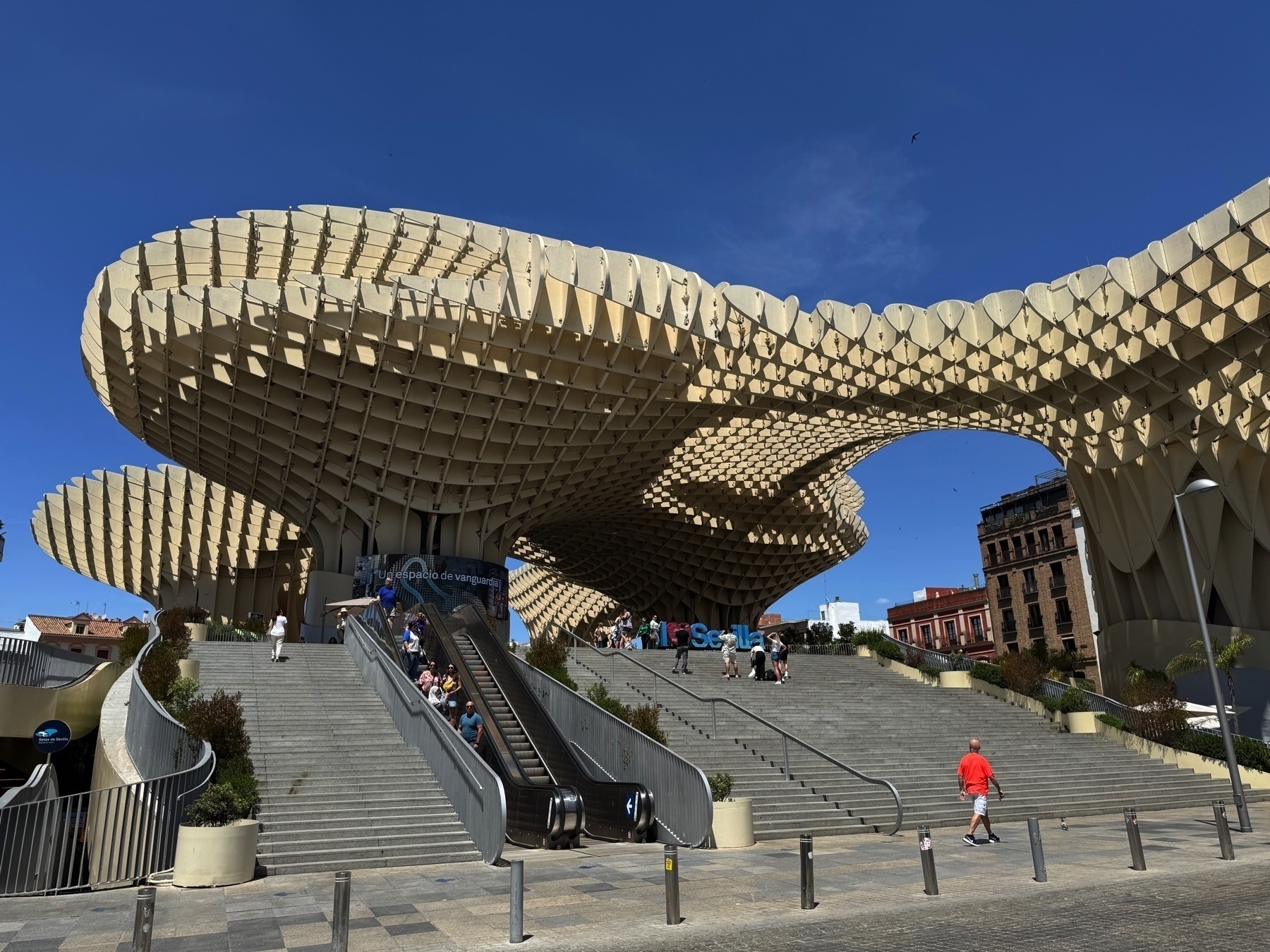 A sweeping view of Metropol Parasol, a massive wooden structure with curving, honeycomb-like forms, rising above a wide plaza with escalators and stairways. People rest on the steps and admire the architectural landmark under a clear blue sky.