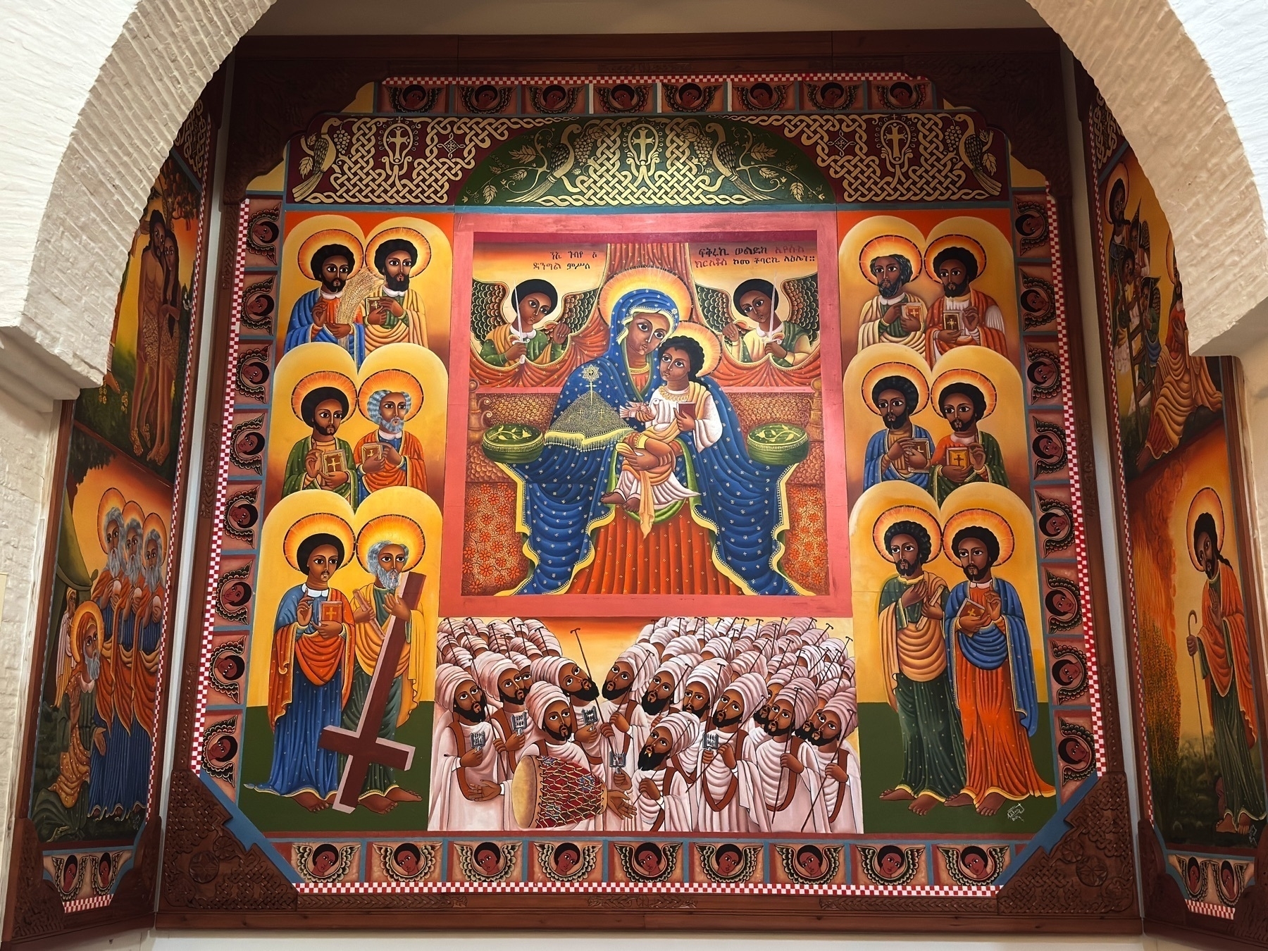 Vibrant religious mural depicting saints and holy figures in stylized, symmetrical arrangements. Rich gold and red tones, along with intricate patterns, highlight the iconographic Ethiopian Christian art style.