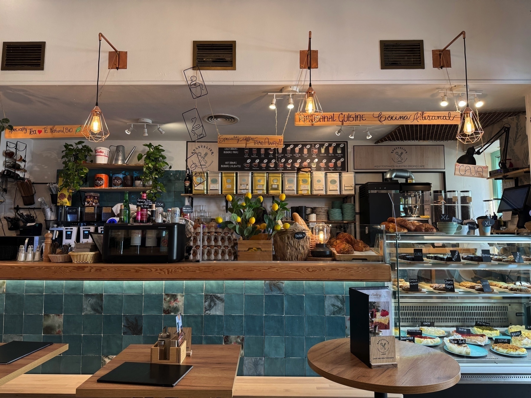 A cozy café interior with teal tiled walls, warm wood accents, and hanging filament bulbs. Behind the counter are pastries, fresh eggs, and coffee-making gear, while a display case on the right showcases cakes and baked goods, creating a charming, artisanal atmosphere.