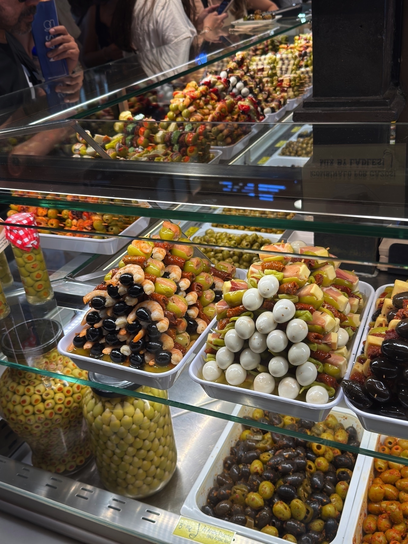 Close-up of a glass display case filled with colorful skewers of marinated olives, cheese, cherry tomatoes, pickles, and cured meats. The skewers are arranged in neat pyramids, with glossy textures and vibrant colors that highlight their freshness and variety. Jars of green and stuffed olives add to the visual appeal, and the reflection of customers adds a dynamic sense of movement.