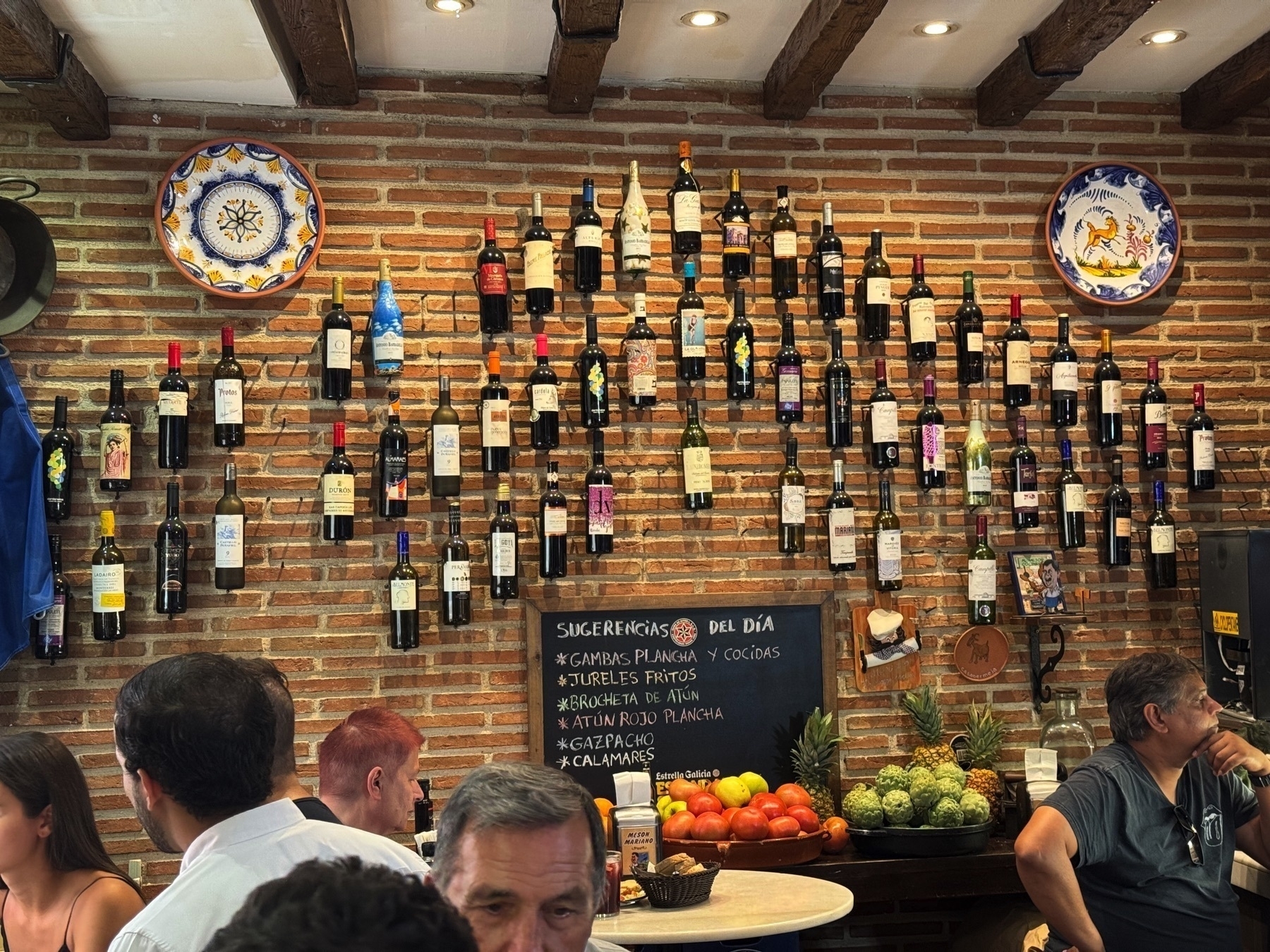 A vibrant interior scene with diners in the foreground and a wall full of wine bottles organized in vertical lines against rustic brick. Colorful ceramic plates and a central chalkboard listing daily specials add charm, while fresh fruit on the counter brings a pop of natural color to the cozy, bustling restaurant atmosphere.