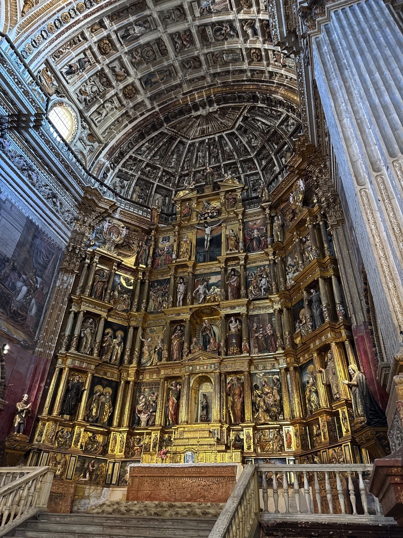 Side-angle view of the richly detailed altarpiece, composed of multiple niches filled with polychrome religious statues and gold accents. The vaulted ceiling above is intricately carved and painted, adding depth and drama to the vertical space.