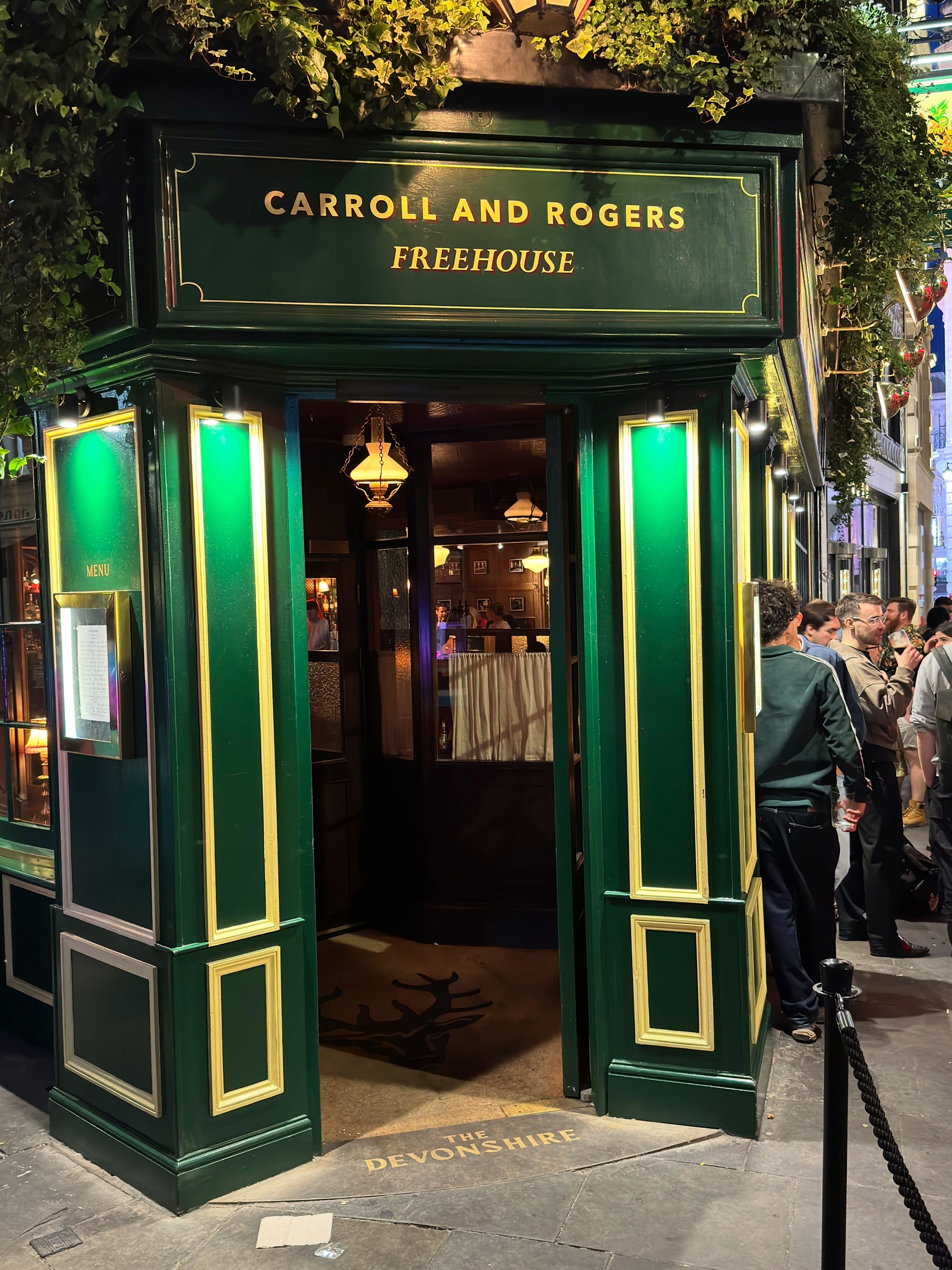 The green and gold entrance of Carroll and Rogers Freehouse stands open to the street, framed by ivy and glowing panels, as groups of people cluster nearby waiting to get inside on a busy night.