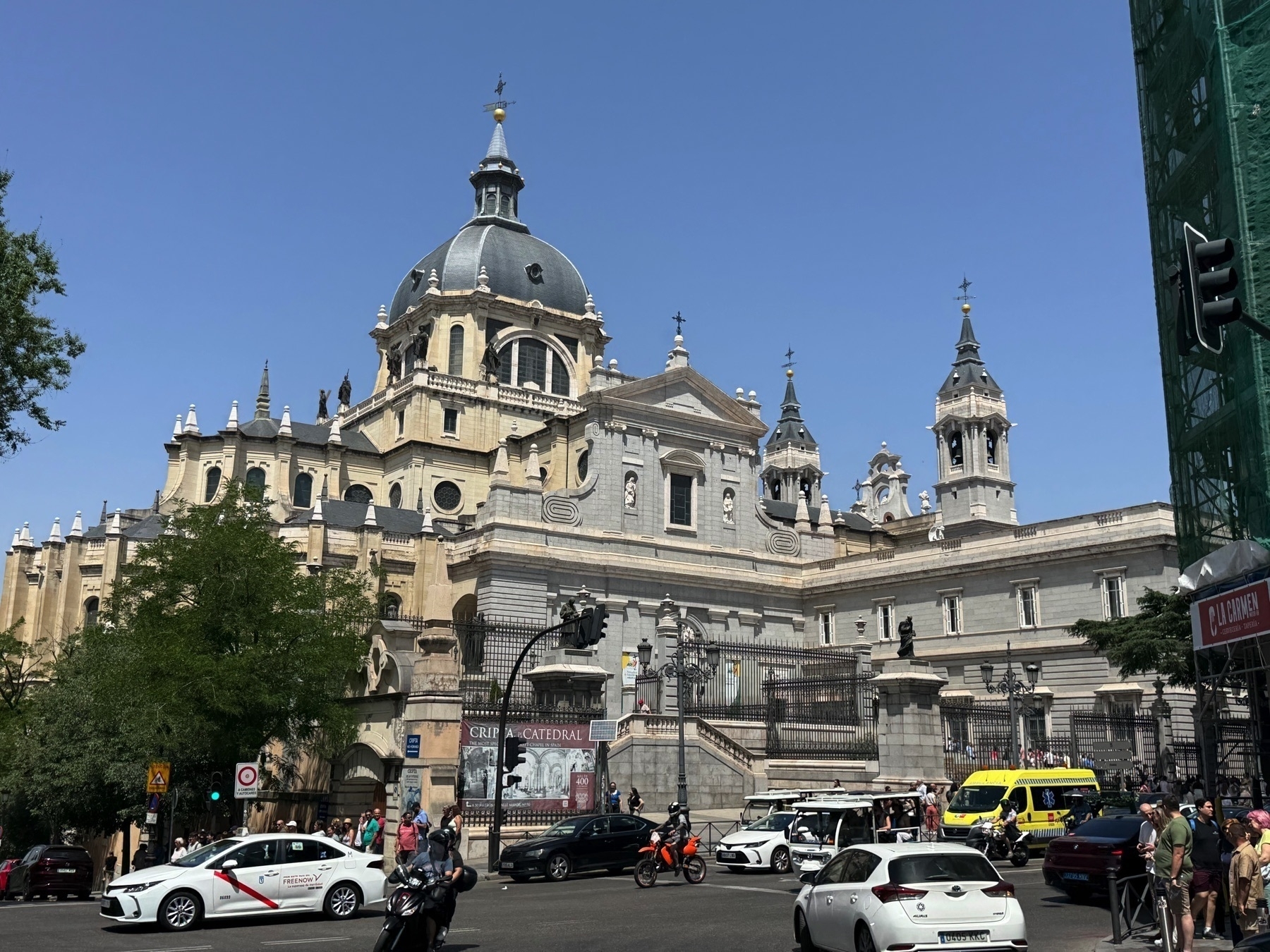 Street scene in front of the Almudena Cathedral with a mix of tourists, vehicles, and historic architecture. The cathedral’s ornate dome and towers rise prominently against a clear blue sky. A steady flow of cars, including a marked taxi and an ambulance, passes along the busy road. Trees and signage frame the scene, capturing the dynamic intersection of city life and monumental history.