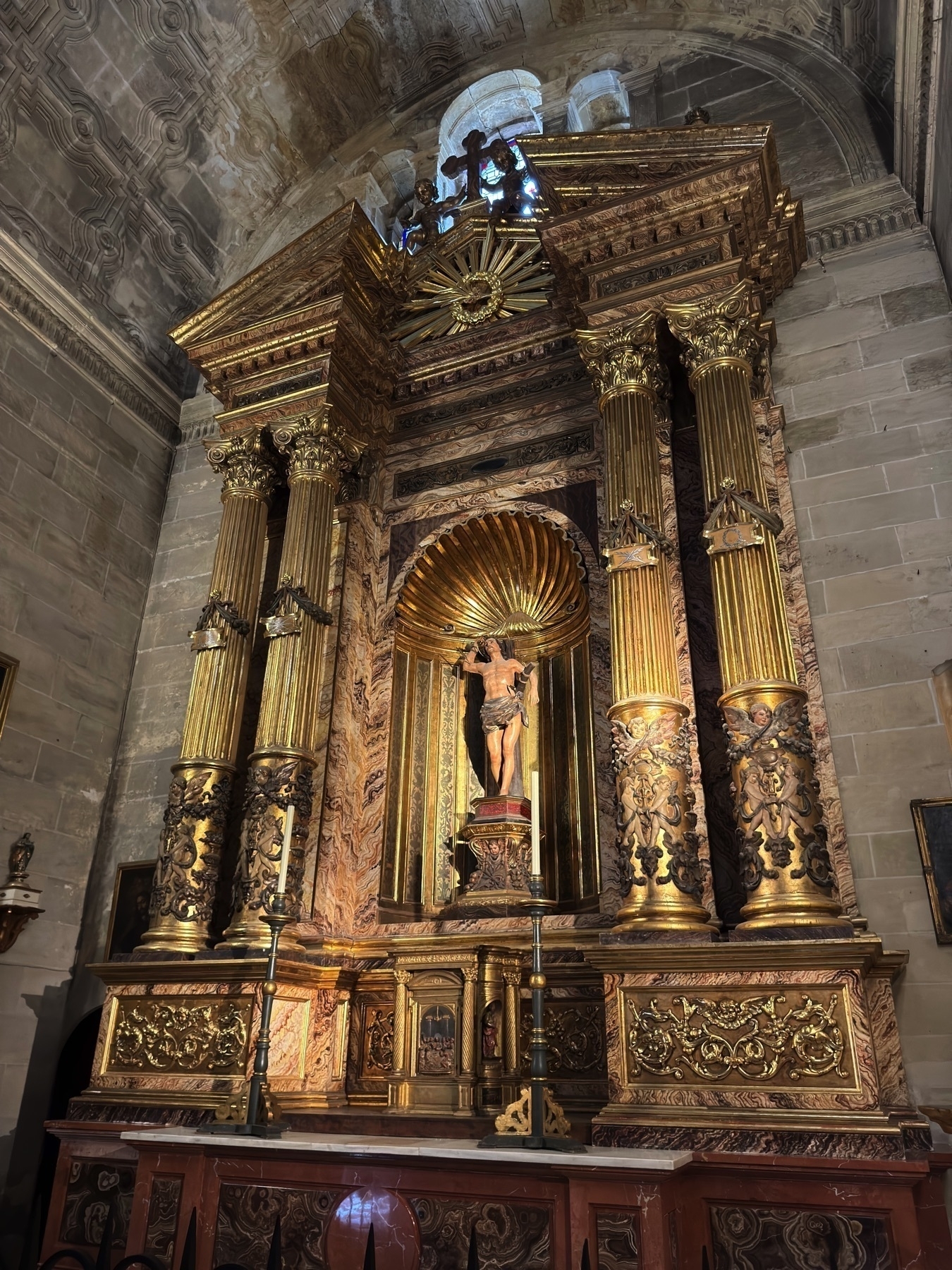 An opulent side altar made of marbled stone and gilded columns, featuring a crucified Christ figure beneath a scalloped golden niche. The surrounding architecture displays a mix of baroque textures, blending deep reds, golds, and intricate patterns.