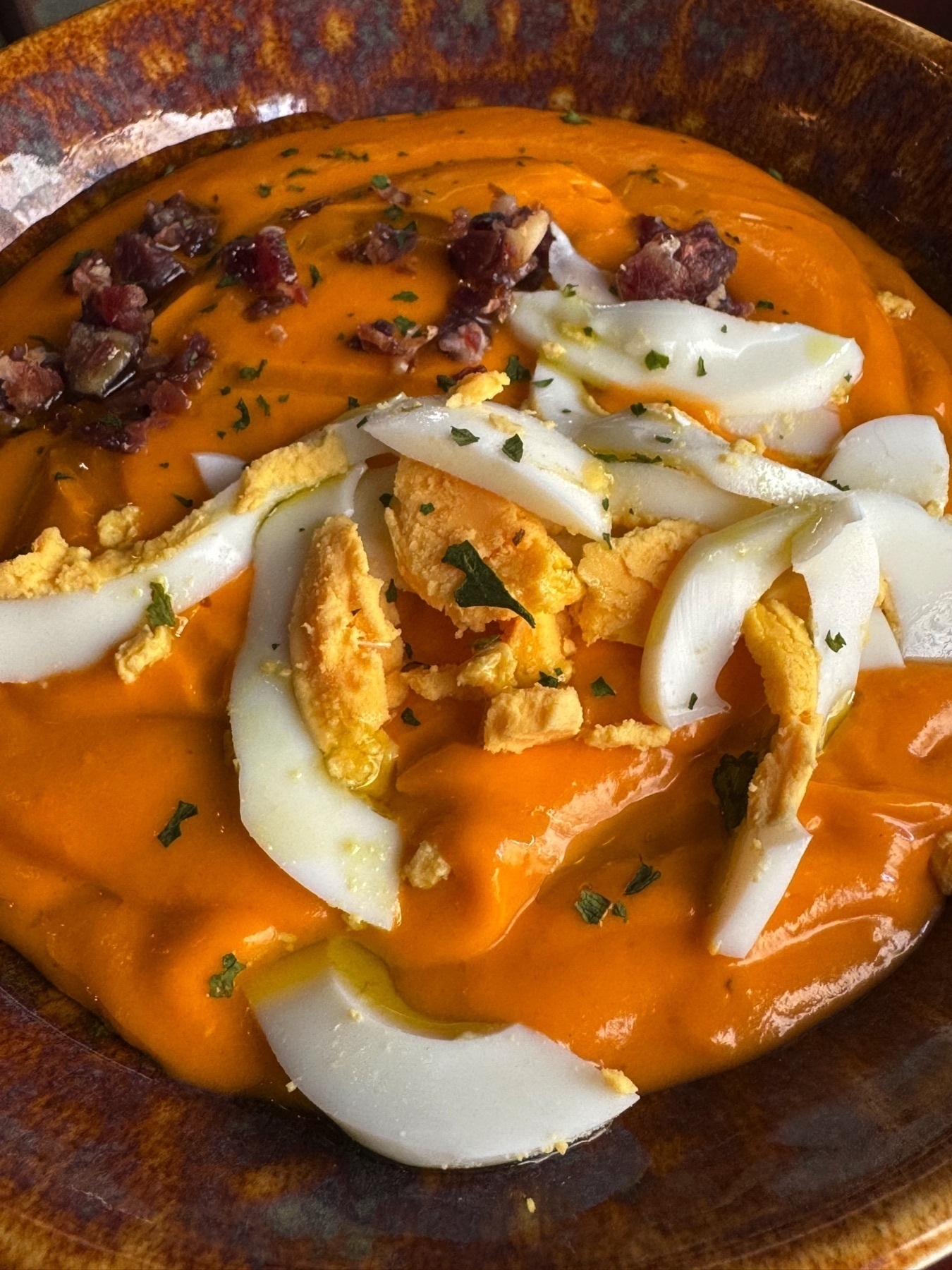 Bowl of creamy orange salmorejo garnished with finely chopped cured ham and slices of hard-boiled egg, offering a rich, velvety texture and savory toppings.