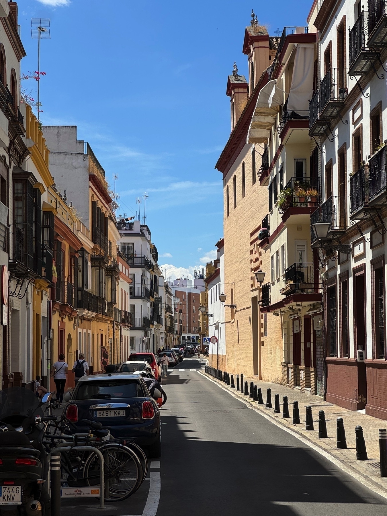 A narrow urban street lined with parked cars and flanked by traditional Spanish buildings painted in warm tones of yellow, red, and white. Decorative iron balconies hold potted plants, and pedestrians walk along the sidewalks. In the distance, modern apartment buildings rise, blending old and new architecture under a bright blue sky.