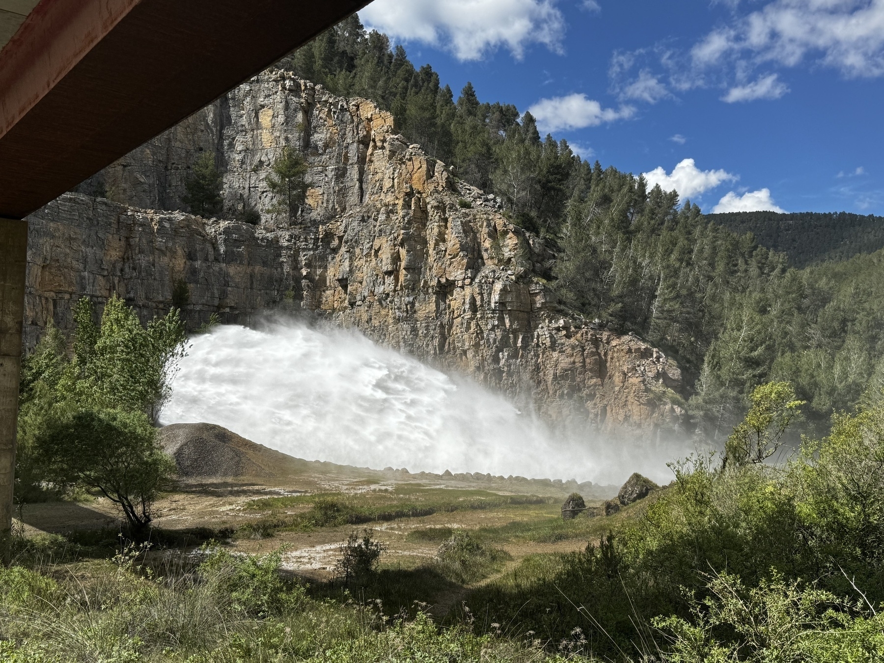 Water powerfully gushing from a dam outlet set in a rocky hillside, surrounded by pine trees and under a bright sky.