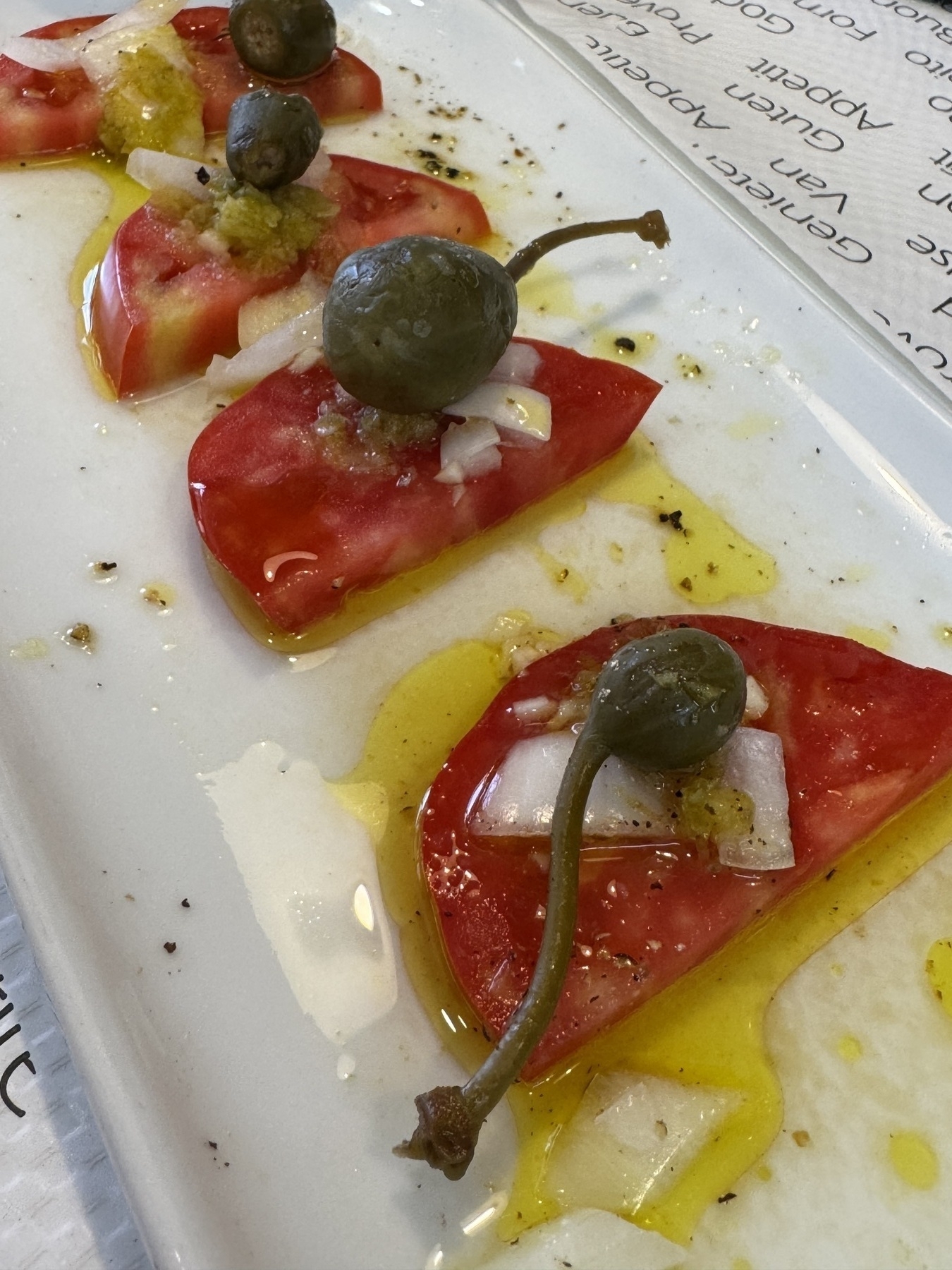 Sliced tomato topped with diced onion, caper berries, olive oil, and seasoning, served as a light starter.