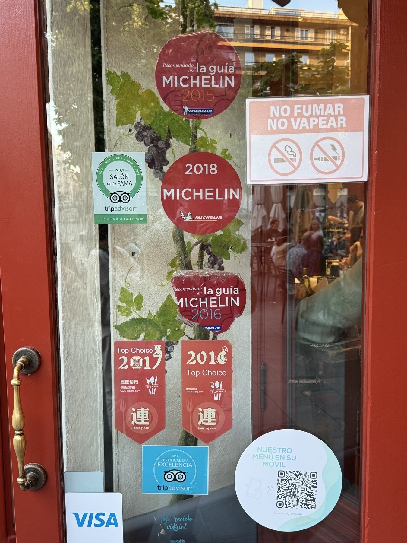 A restaurant door with multiple accolades including Michelin Guide recommendations for 2015, 2016, and 2018, and TripAdvisor “Certificate of Excellence” and “Hall of Fame” awards. The window also displays “Top Choice” recognitions in Spanish and Chinese, a grapevine graphic, and a QR code for the digital menu.