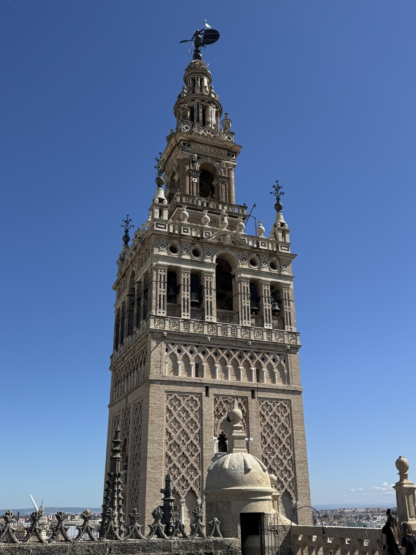 Striking view of La Giralda’s ornate upper levels, featuring detailed brickwork, Moorish arches, and the bronze weather vane known as El Giraldillo silhouetted against a vibrant blue sky.