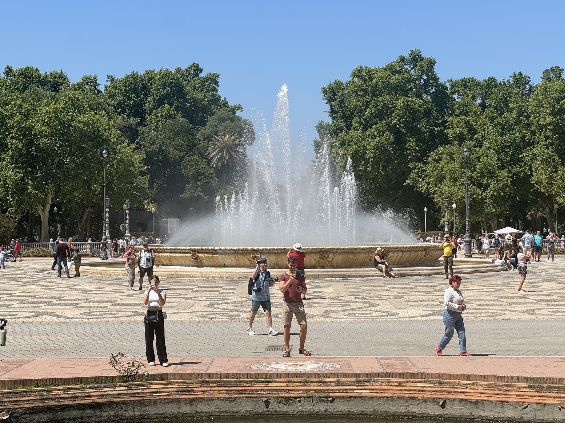 Detailed view of the large circular fountain, with streams of water arching high into the air. Visitors rest along the edge, walk nearby, or take photos. The patterned stonework and lush trees around the plaza create a tranquil, vibrant scene.
