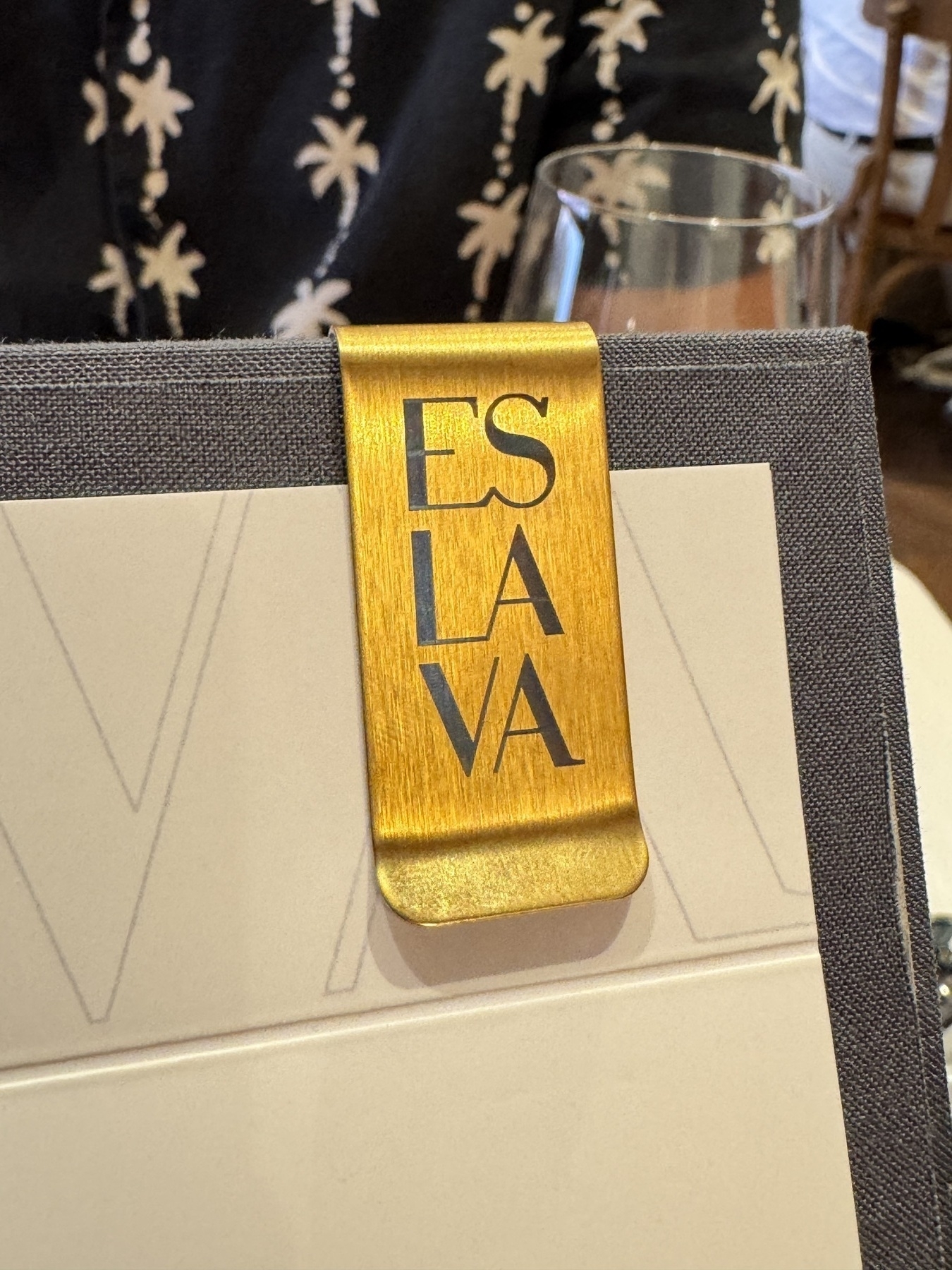 A close-up of Eslava’s menu, featuring a textured gray cover held by a shiny gold clip engraved with the restaurant’s name, with a wine glass and patterned shirt visible in the softly blurred background.