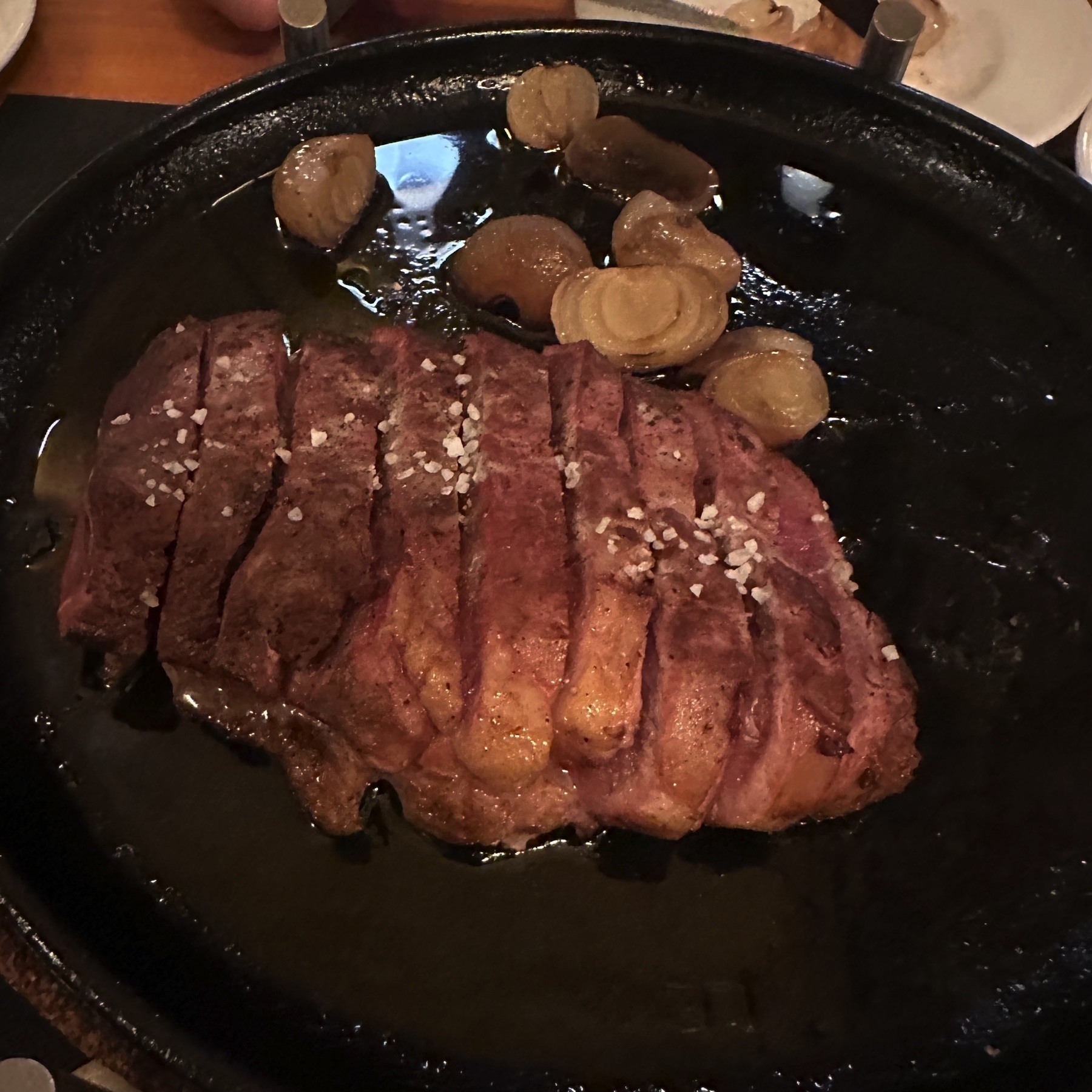 A sizzling cast-iron platter with thick slices of grilled duck, seared to a deep golden-brown and topped with coarse sea salt. Surrounding the meat are roasted shallots glistening in a rich layer of pan juices, creating a rustic and hearty presentation that emphasizes the meat’s tenderness and flavor.