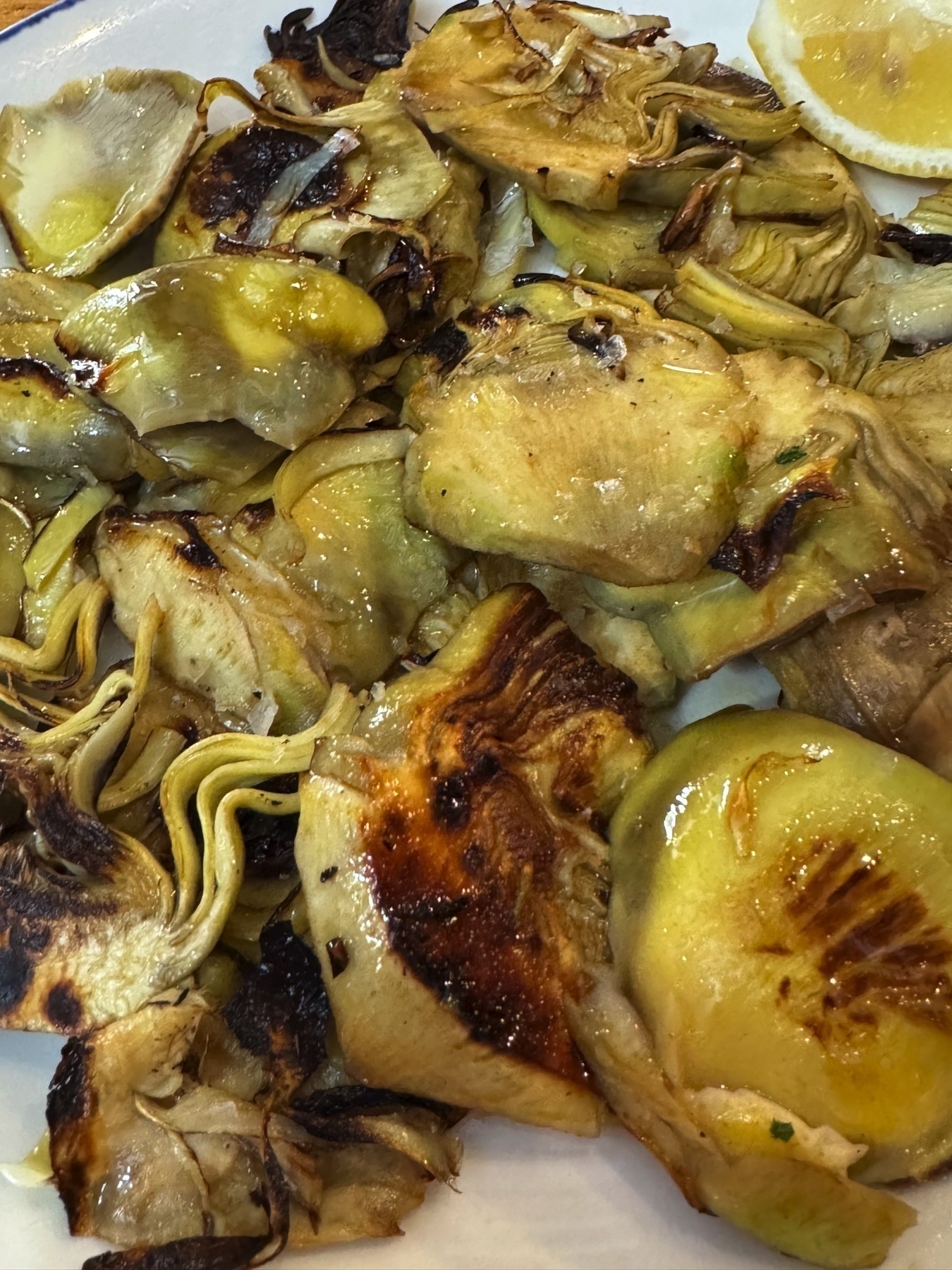 A plate of grilled artichokes sliced thin, beautifully seared to a golden brown with slightly crispy edges and tender centers, glistening with olive oil and served with a wedge of lemon for brightness. The rustic presentation emphasizes the earthy, rich flavor of the artichokes.