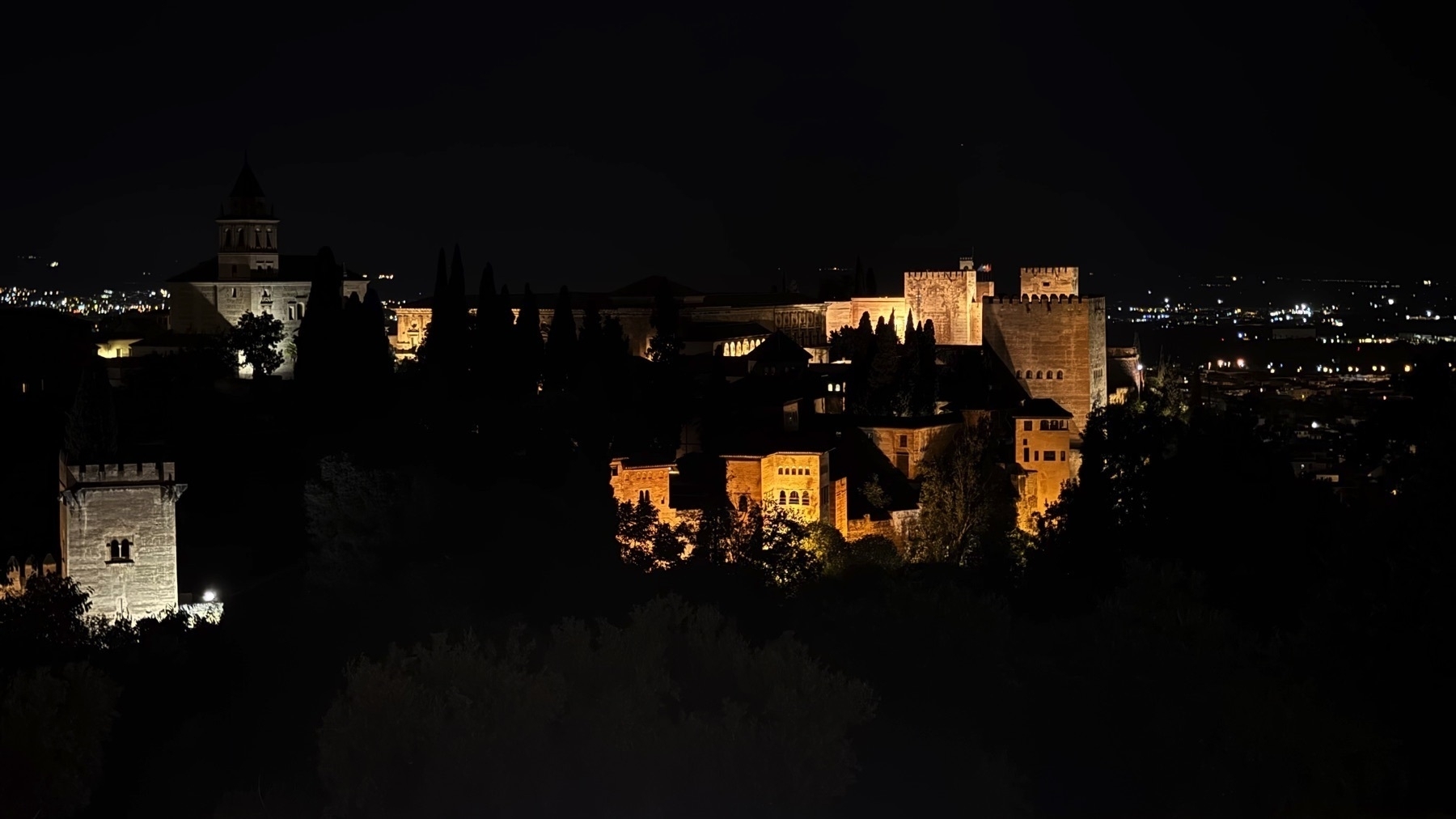 Nighttime view of the Alhambra complex from a distance, with its iconic towers and palace structures—such as the Alcazaba and Palacio de Carlos V—bathed in golden light against the dark hillside.