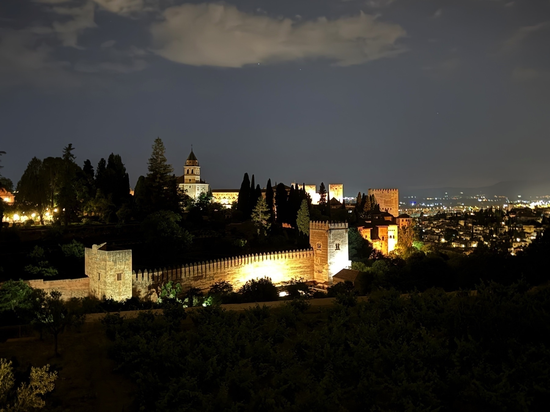 Panoramic nighttime view of the Alhambra complex from a distance, with its illuminated towers and walls—including the Torre de Comares and Torre de la Vela—rising above treetops, and the lights of Granada city twinkling in the background.