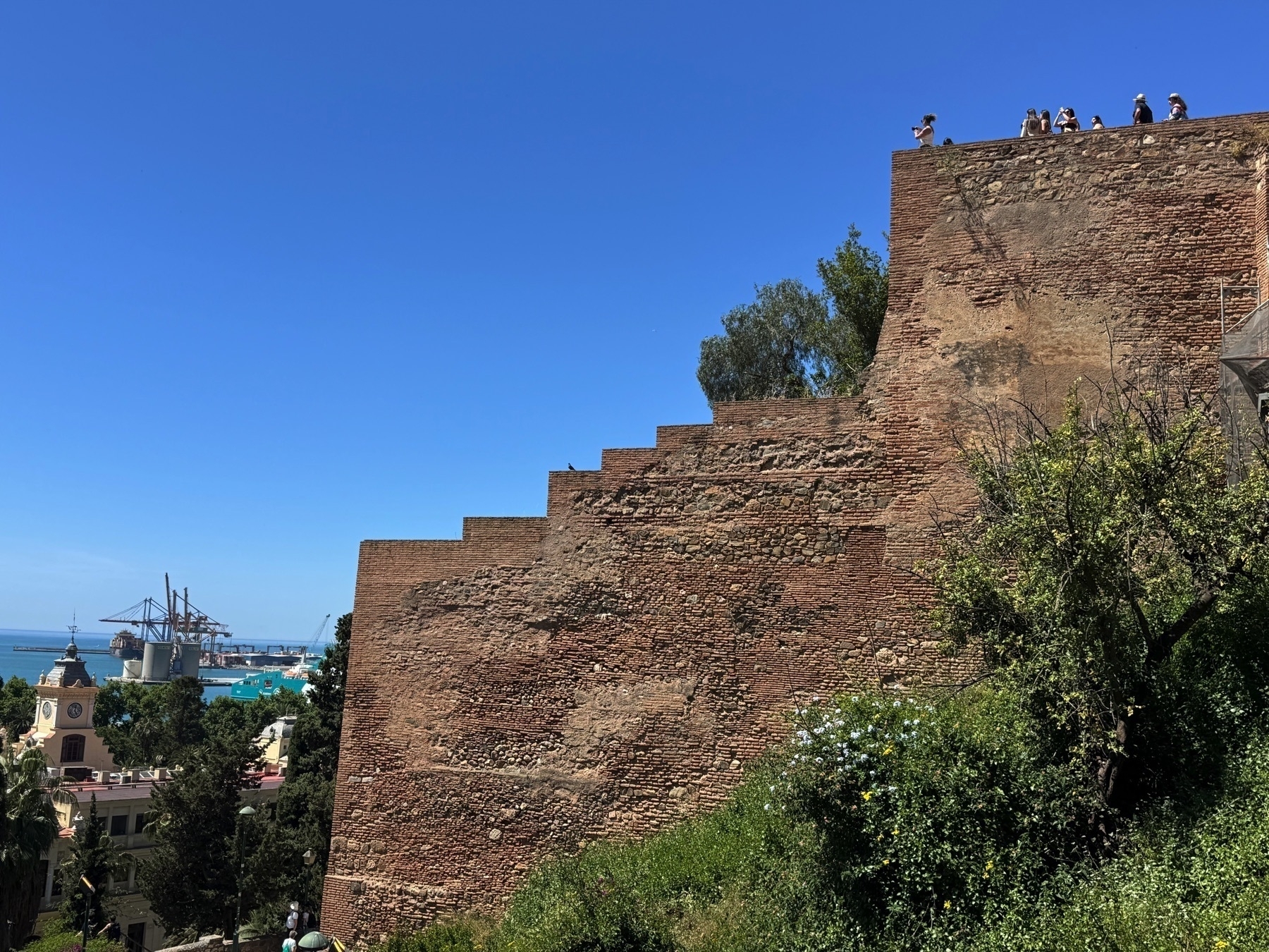 View of the stepped brick walls of the Alcazaba of Málaga descending toward the city. Tourists stand atop the fortress, with the modern port and container cranes visible beyond, under a deep blue sky.