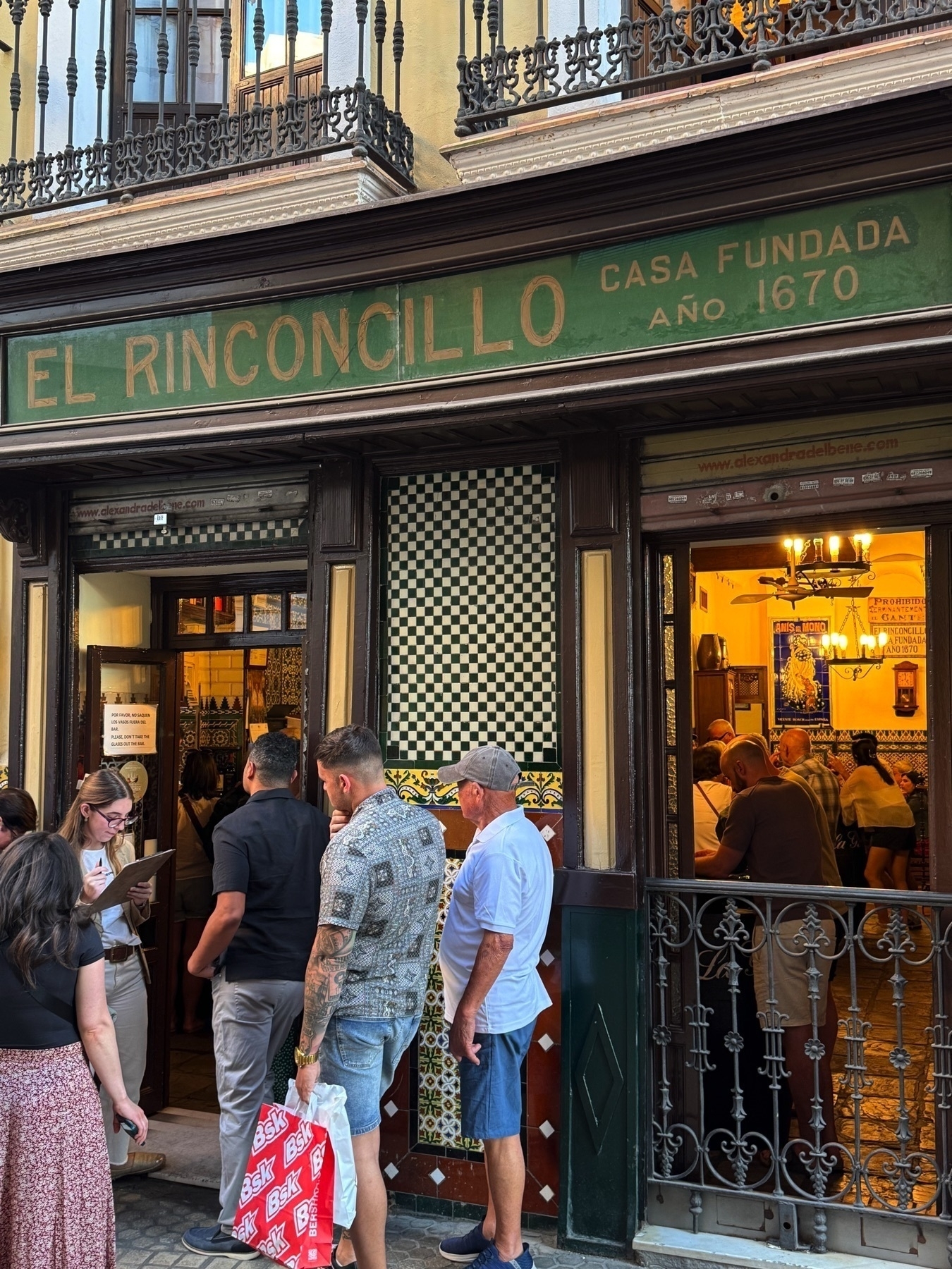 Street-level view of the historic El Rinconcillo restaurant with its green sign, tiled facade, and crowd of people waiting to enter, conveying the popularity and old-world charm of the establishment.
