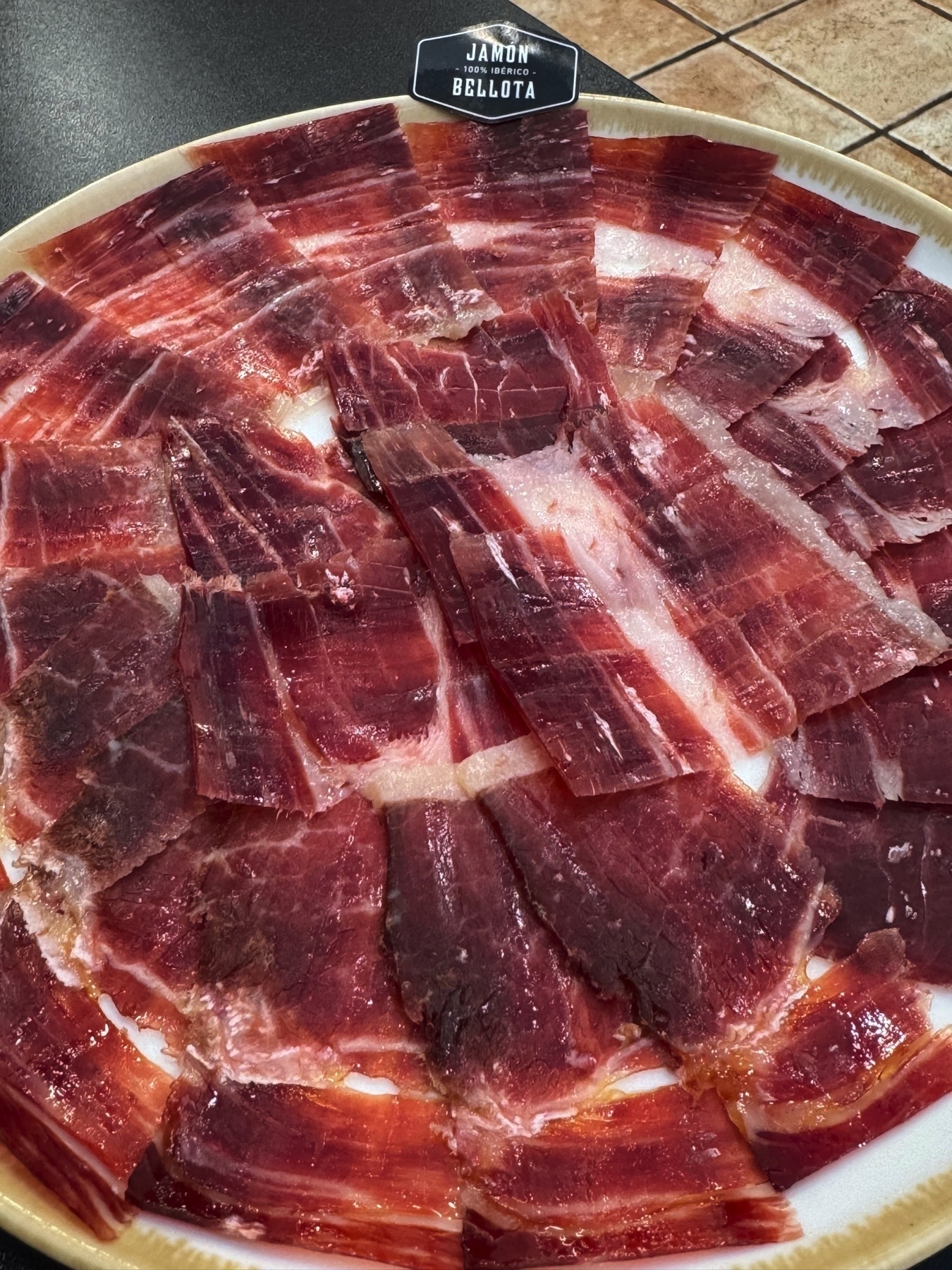 A plate of finely sliced Jamón Ibérico de Bellota, arranged in a circular pattern to showcase the marbling and deep reddish hues of the cured ham. The translucent slices reveal streaks of intramuscular fat, indicating the high quality of the meat. The glossy surface and delicate texture make it look rich and savory, with a small sign at the top of the plate labeling it as “Jamón 100% Ibérico Bellota.”