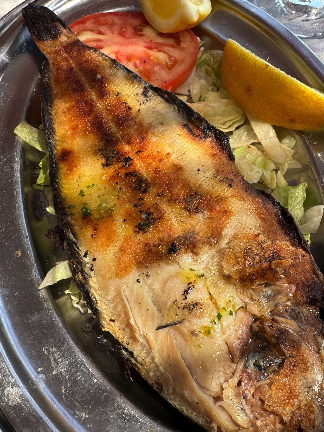 Chargrilled fish fillet served on a metal tray atop shredded lettuce and tomato slices. The fish has a crispy, golden skin and is garnished with herbs and lemon wedges.